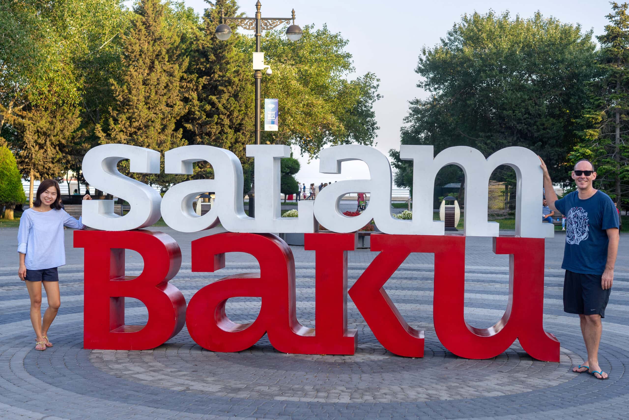 We are standing next to a red and white sign saying Salam Baku on the Bulvar in the Azerbaijan Capital before traveling overland to Yerevan
