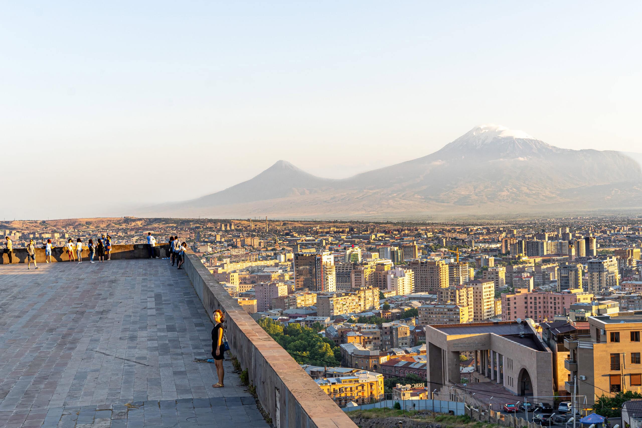Jin standing on a lookout point in Yerevan with the city below her and the snow capped mount Ararat rising on the horizon in the late afternoon light