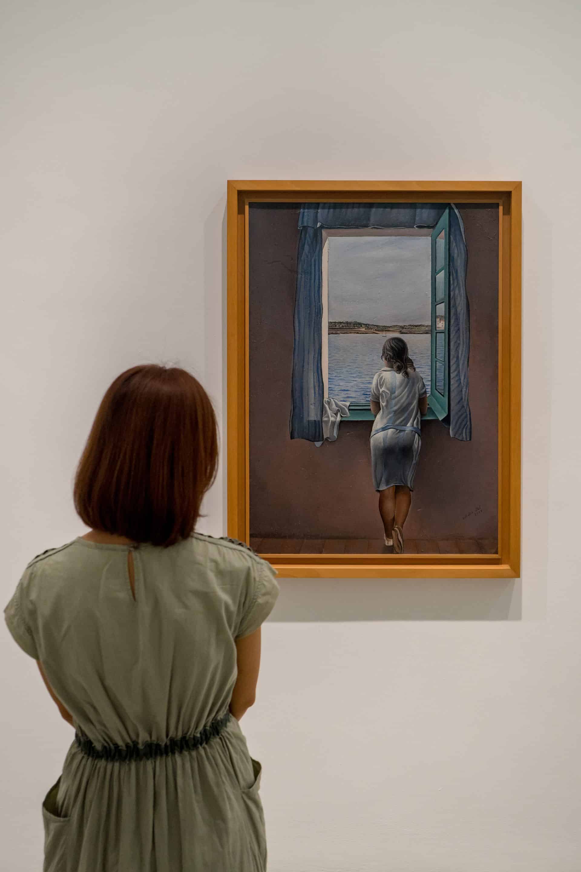 Jin admires a painting in the Reina Sofia. She is standing with her back to camera, looking at a painting of a woman, seen from the back, looking out the window