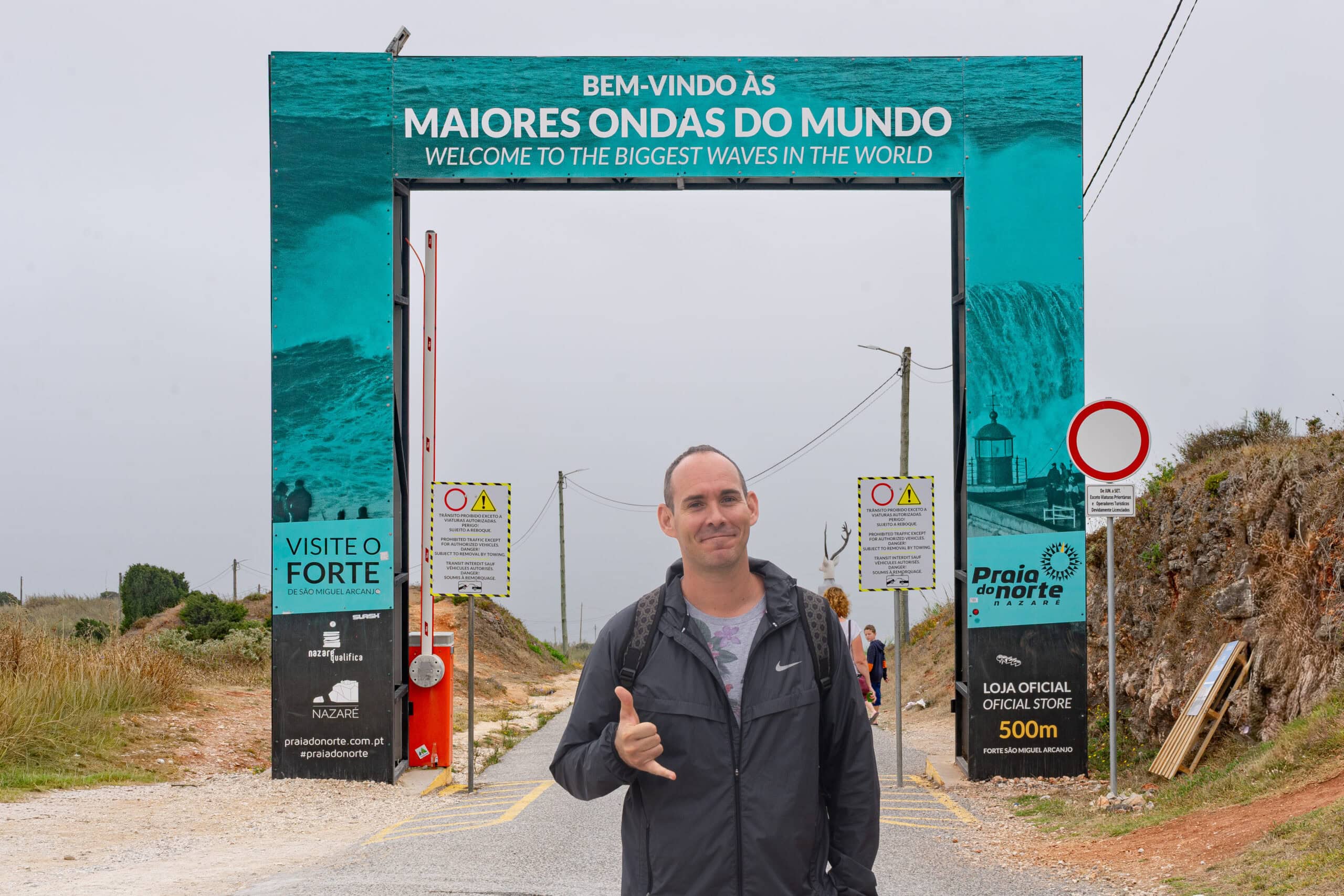 I am standing in front of the entrance to Praia do Norte in Nazare. A blue flat arch behind me says Welcome to the Biggest Waves in the world, written in Portuguese and English