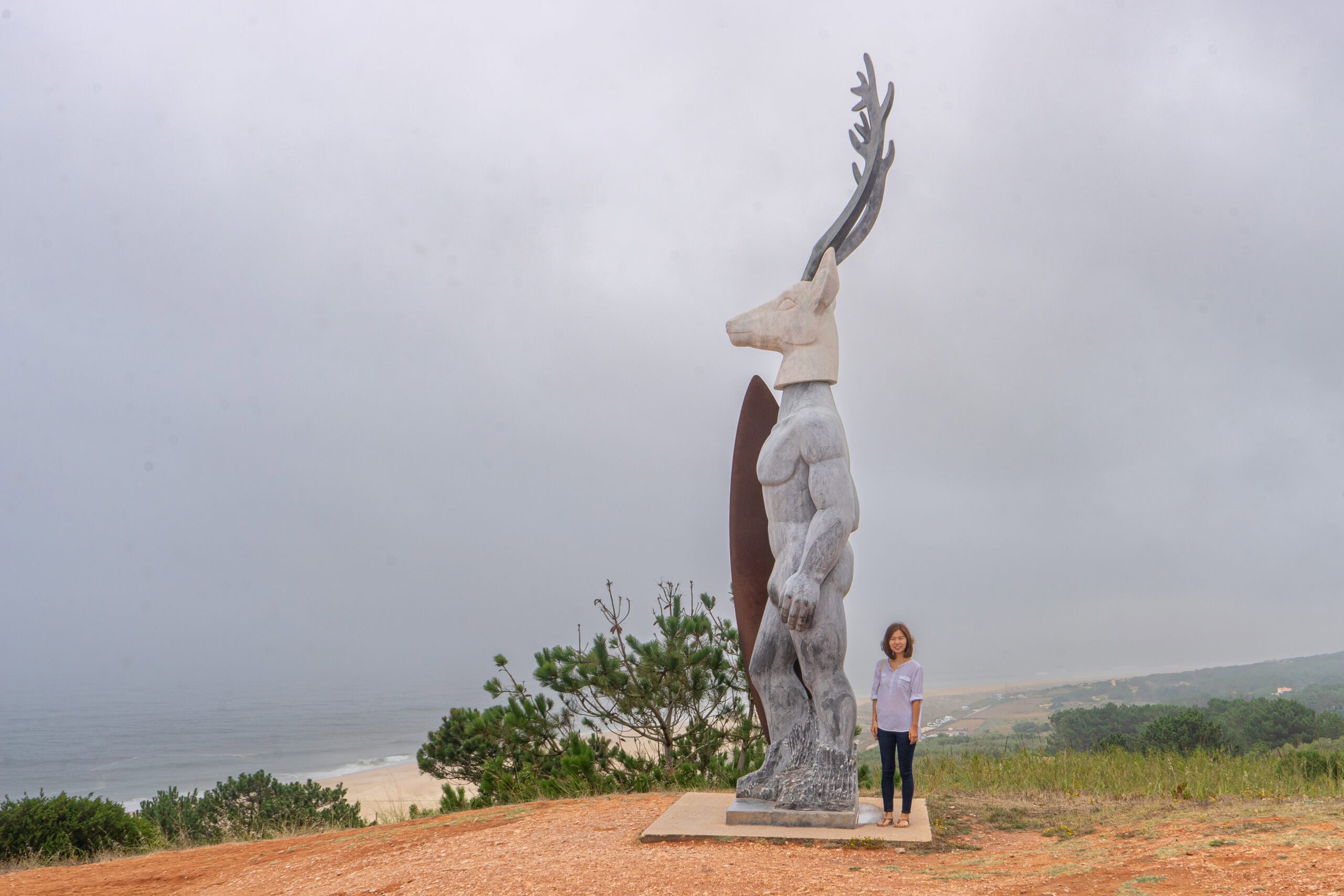 Jin stands next to the statue of Veado. The large statue is about three times her height and depicts a surfer with a deer head. You can see Praia do Norte in the back ground