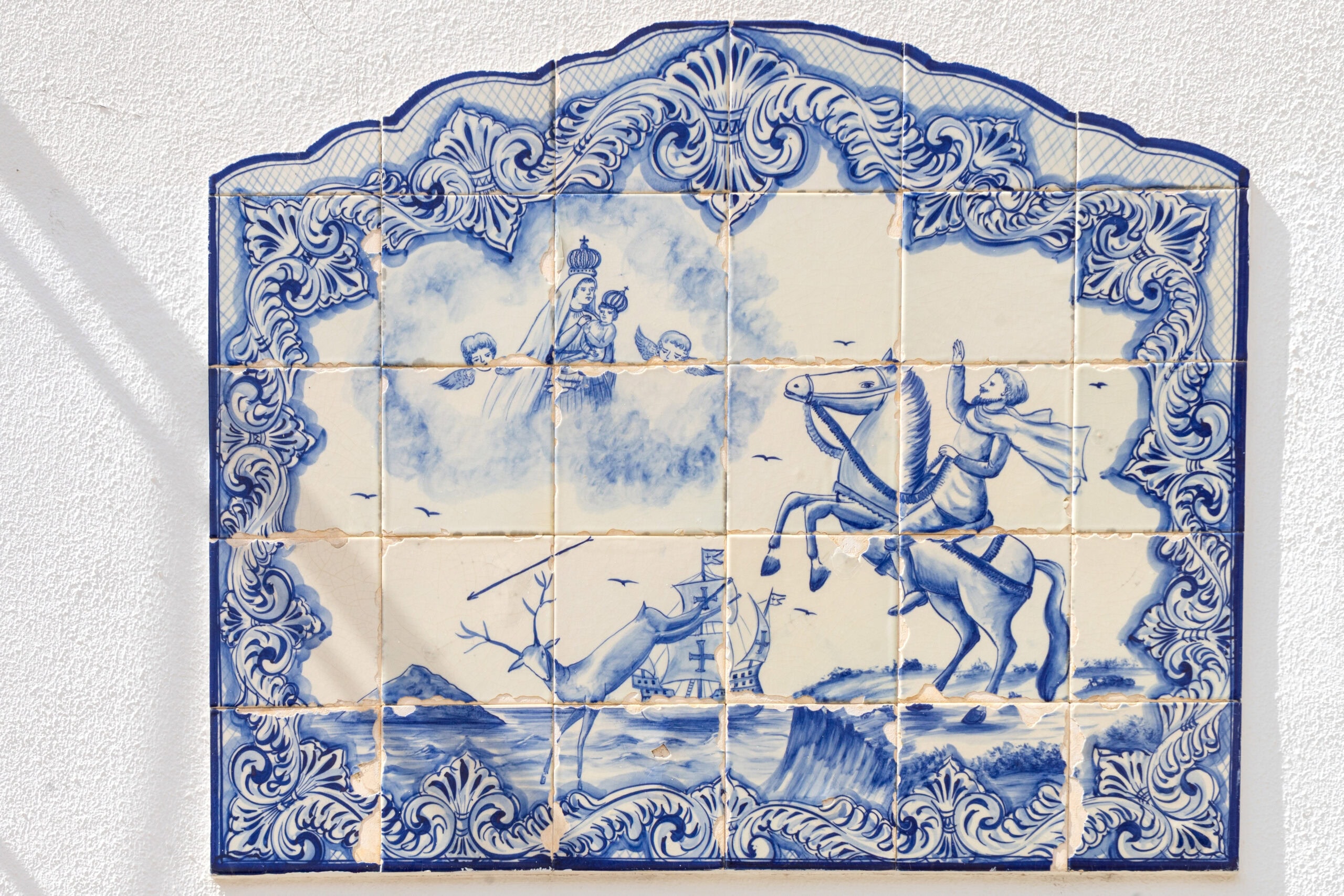 Blue and white Azulejo tiles depicting the Legend of Nazare, seen on a building in Nazare, Portugal. It shows a knight on horseback asking for help from Our Lady of Nazaré, while a deer falls over a cliff to its death 