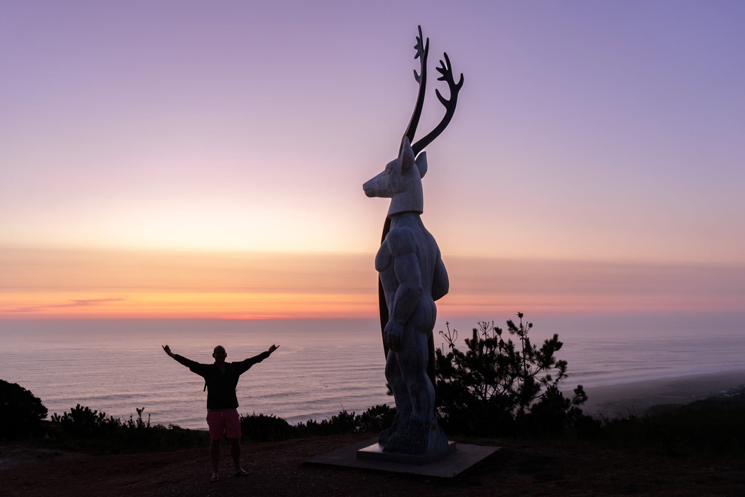 I am posing with my hands in the air next the statue of Veado  at sunset. The sky is warm purple and yellow, with just my silhouette against the Atlantic Ocean behind me