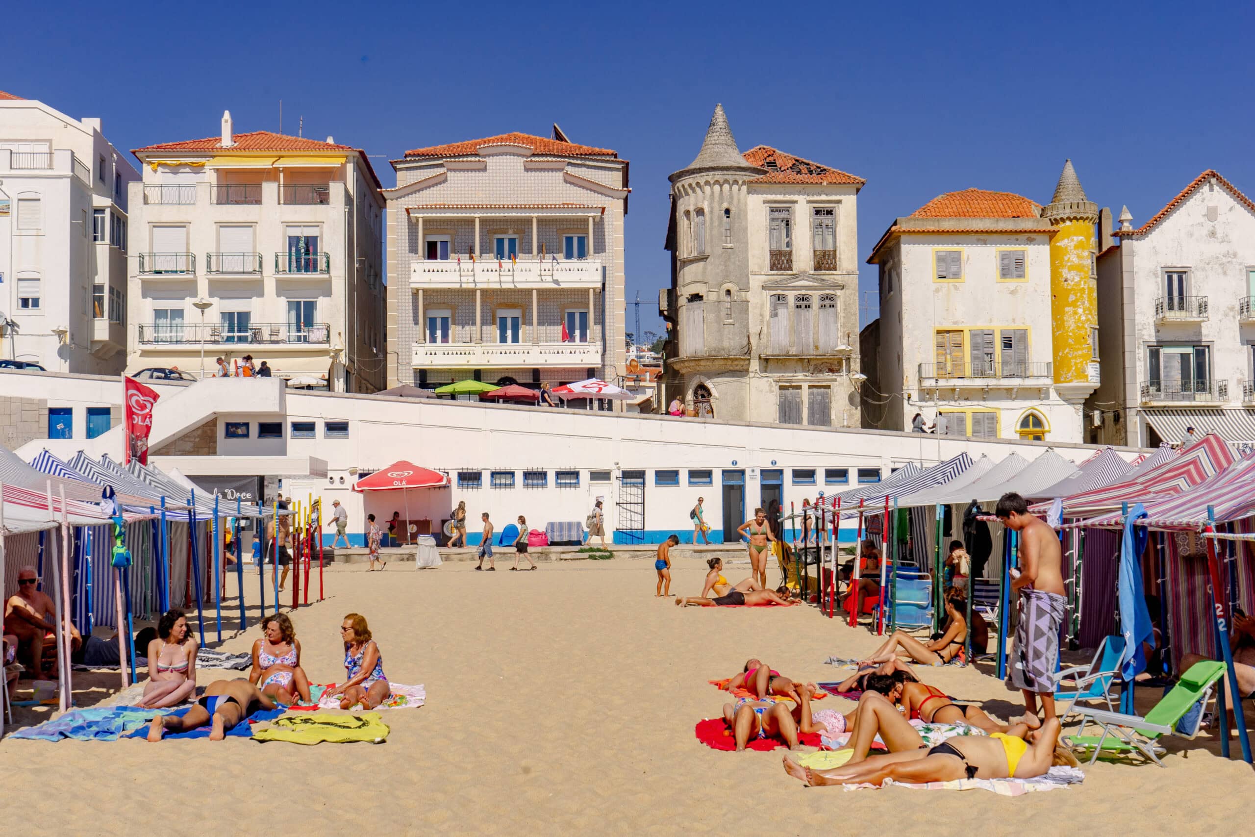 Families sun tanning on the sand next to stripped fabric beach cabanas on Nazare Beach. Just behind the sand you can see houses on the promenade