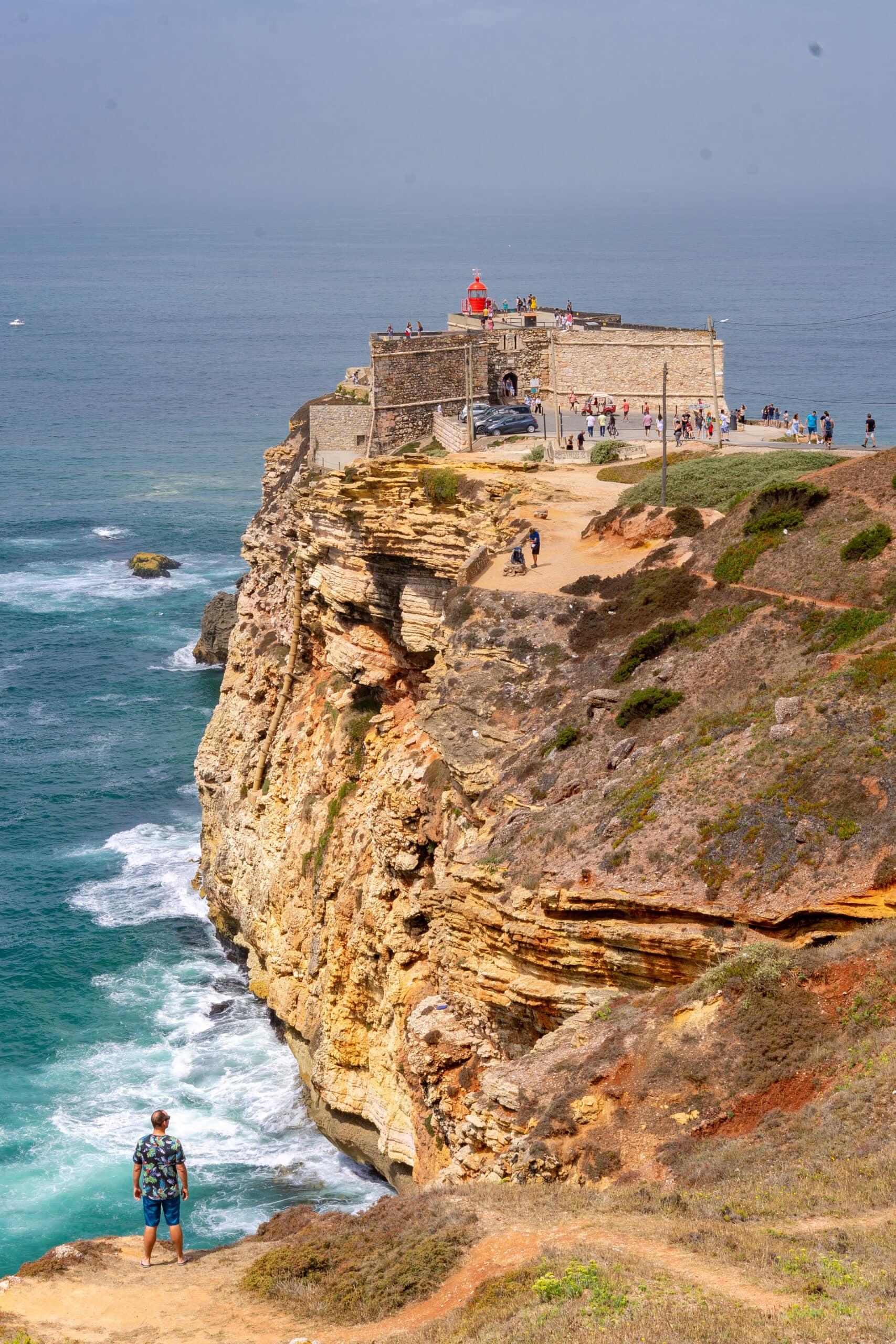 I am standing on a cliff with my back to the camera, looking toward the Nazare Lighthouse