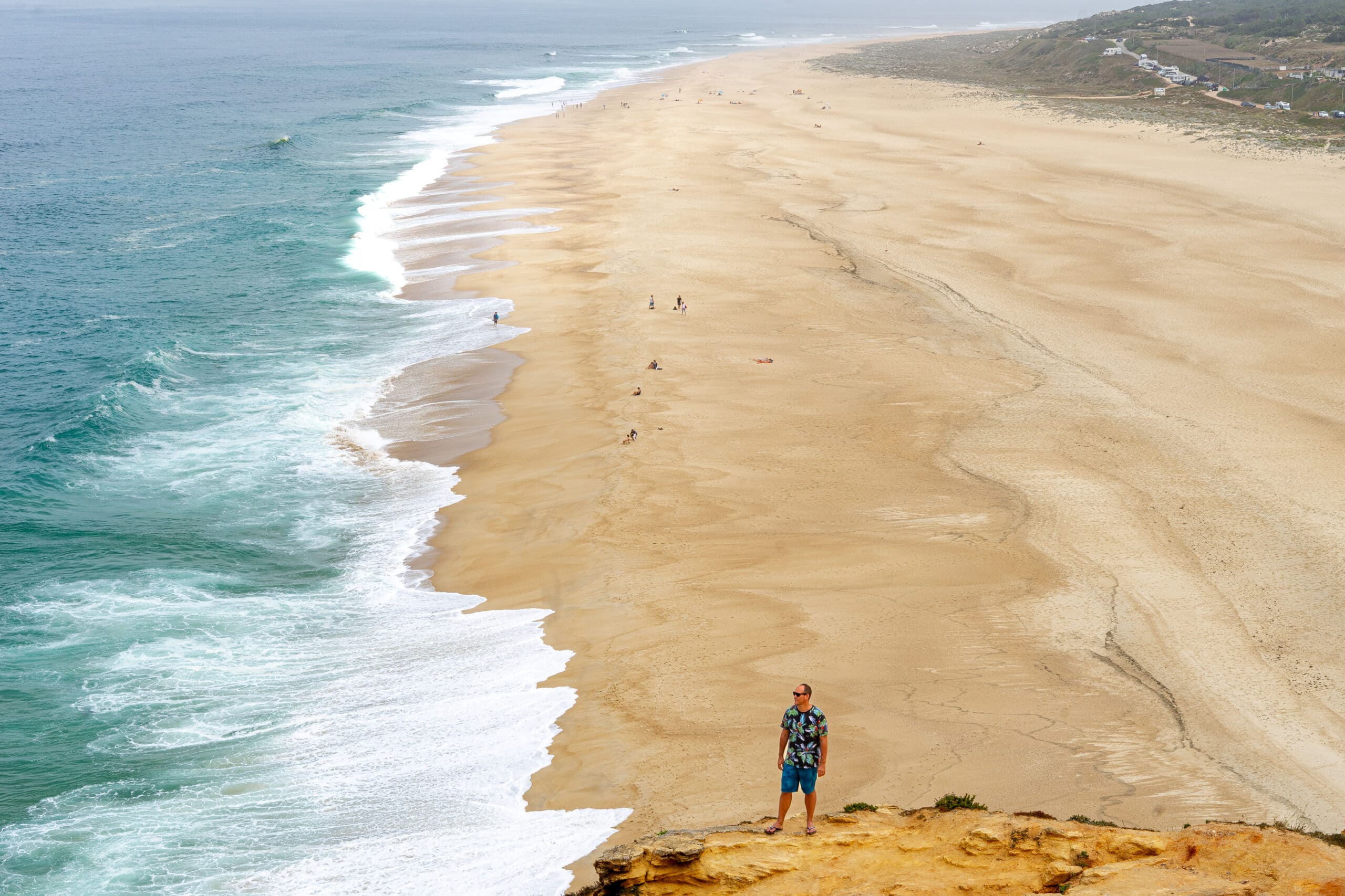 I am standing on a cliff with Praia do Norte behind me. It's a wide, sandy beach with frothy white surf and yellow-brown sand