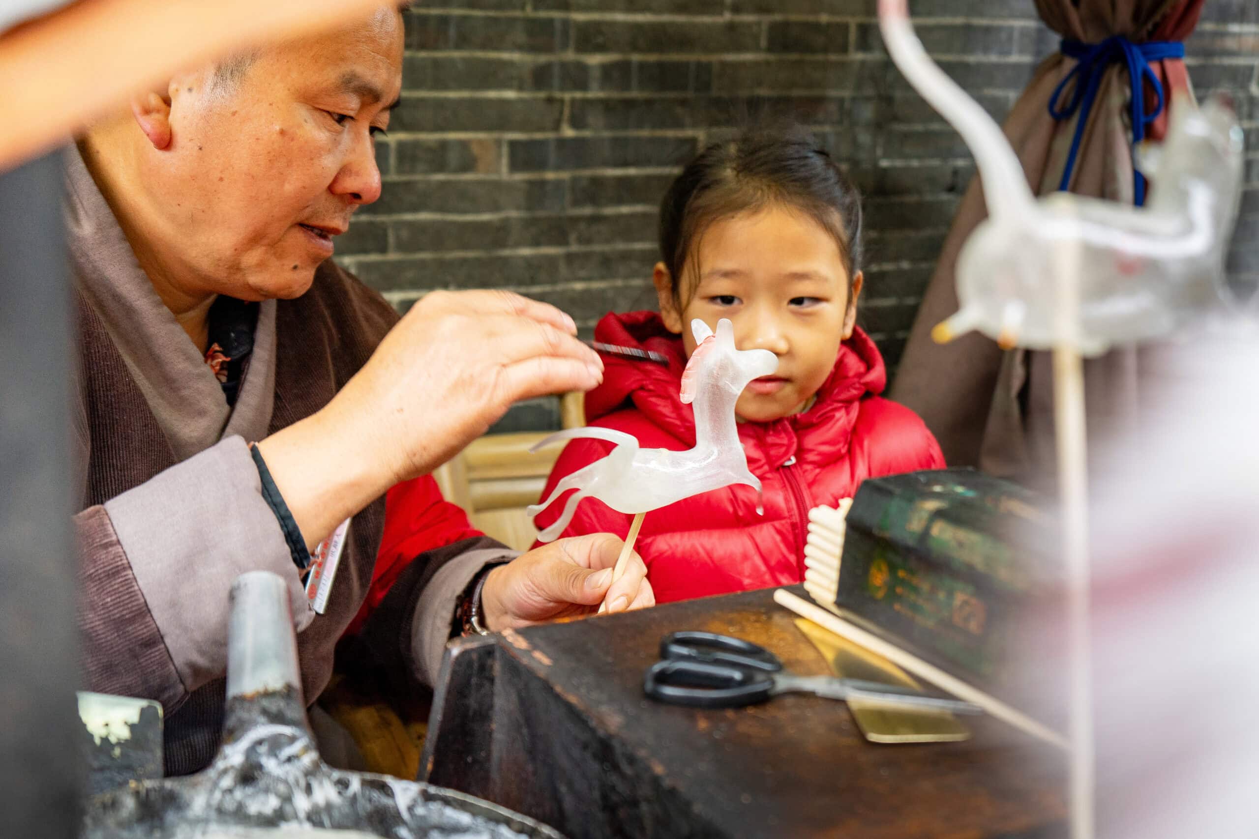 A man creates a horse made from sugar while a young girl watches on eagerly. Jinli Street, Chengdu