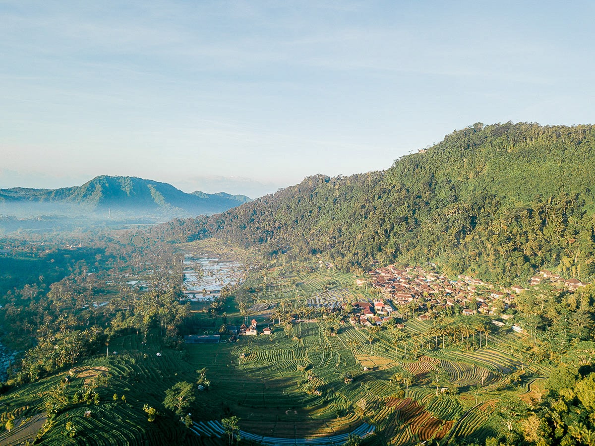 View over Sidemen, with a small town with terracotta roof tiles. The town is surrounded by green forests to one side and rice paddies on the other. There is a mountain in the background