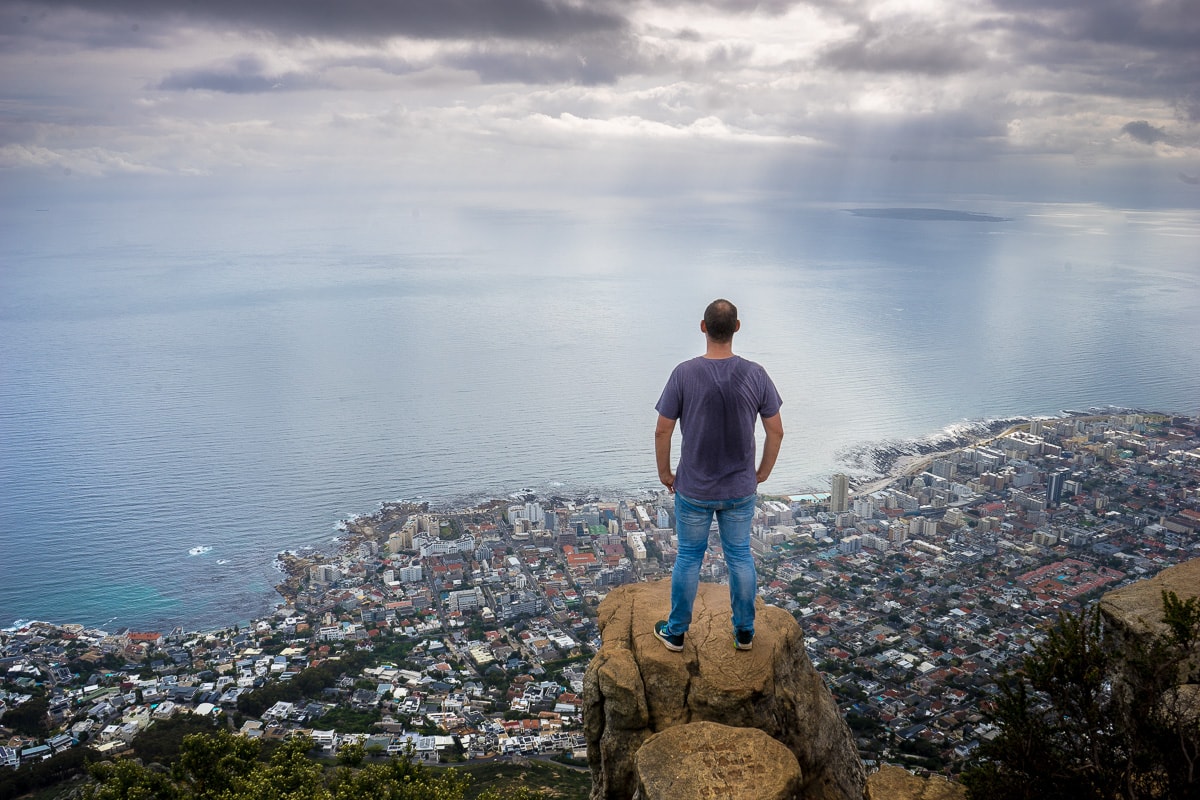 Lion's Head Hike: A man stands on the edge of Lion's Head overlooking the suburb of Sea Point and the Atlantic Ocean. You can see Robben Island in the ocean