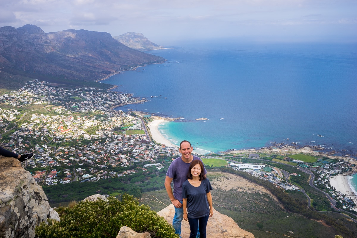 a couple poses for a phot on the summit of Lion's Head with Camps Bay and the Atlantic Ocean in the background 