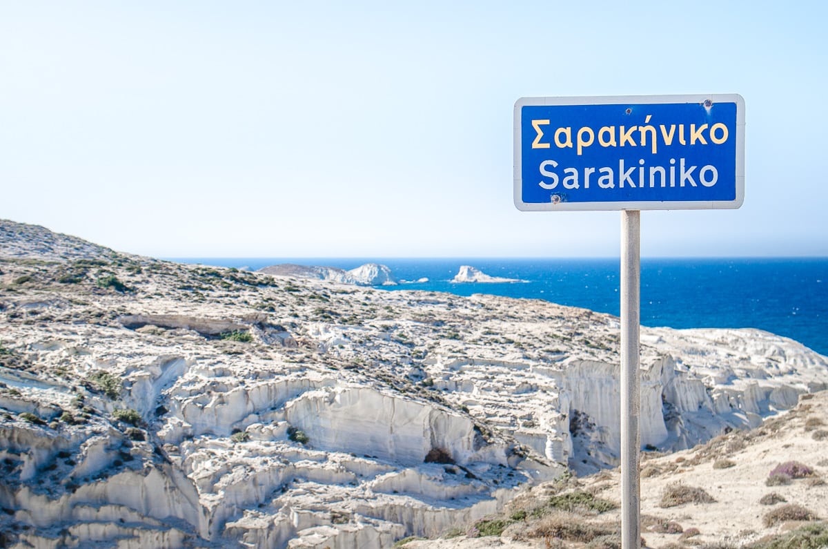 A roadside sign to Sarakiniko Beach on Milos island in Greek and English
