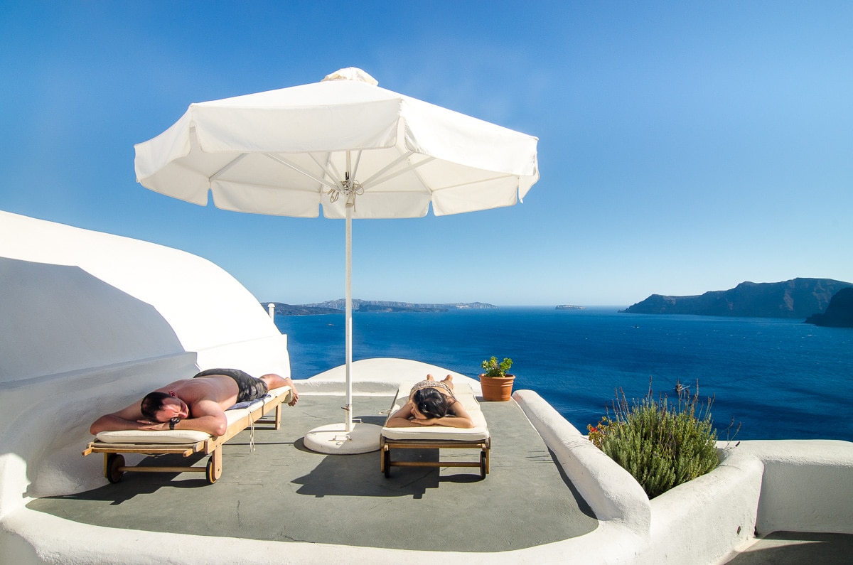 Couple sun bathing under a white parasol with the Santorini caldera in the background