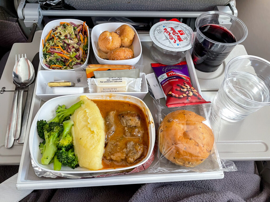 A photo of an economy class meal on Emirates. There's a bread roll, a bag of nuts, a plate with salad, beef in sauce with mashed potatoes and broccoli, cream puffs, water and a glass of wine.  Emirates menu