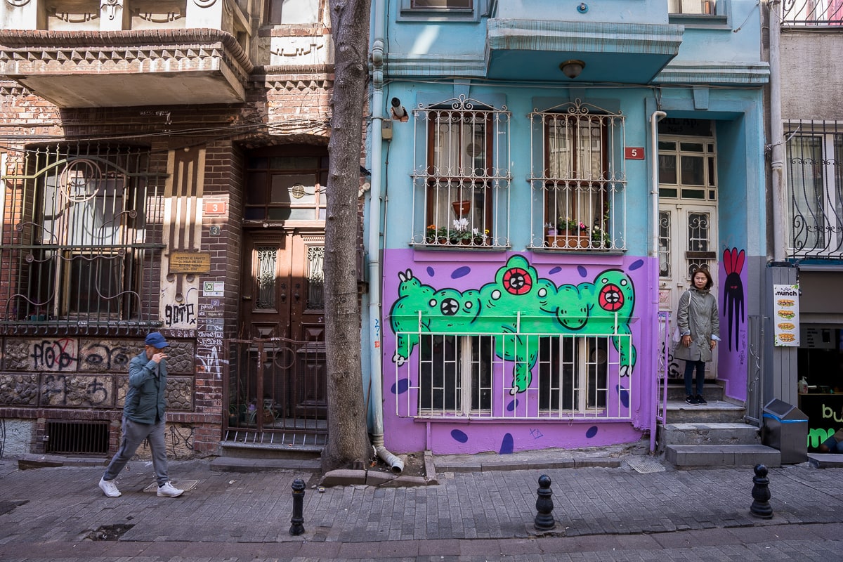 Jin stands in front of a residentail building covered in green and purple graffiti in Kadiköy, Istanbul