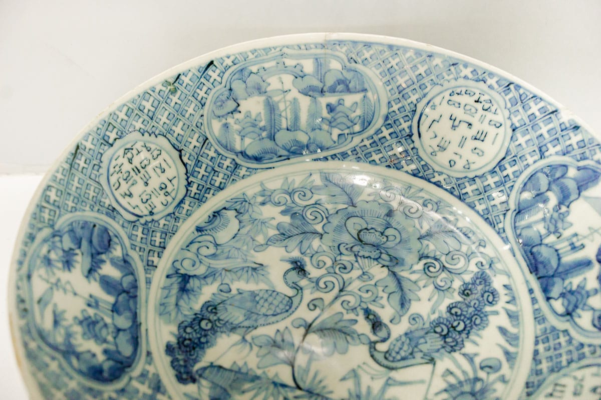 A blue and white Chinese porcelain plate decorated with peacocks and flowers. Surrounding the birds is a trim of geometric patterns. On the trim you can see medallions with Arabic characters 