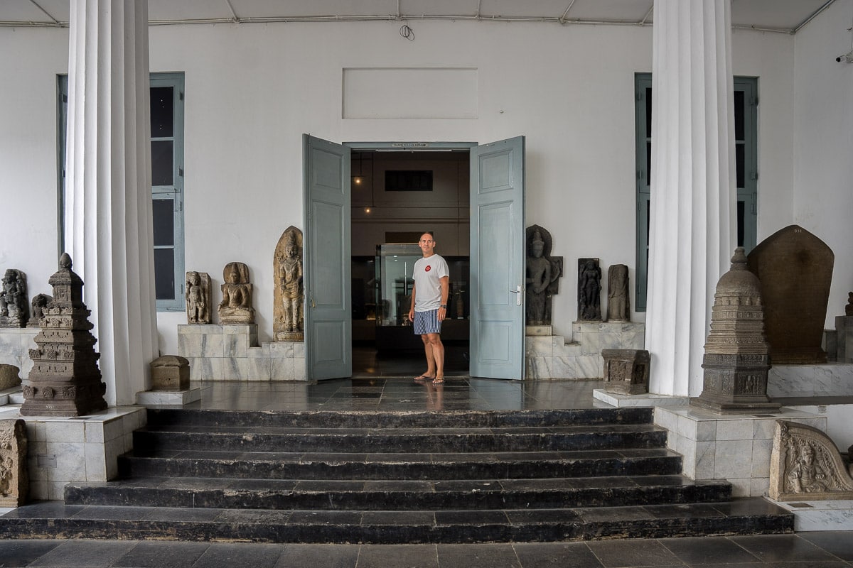 A photo of myself standing between statues with an open doorbehind me leading to the exhibits in the National Museum Indonesia. I'm wearing a white T-shirt and blue shorts with flip flops