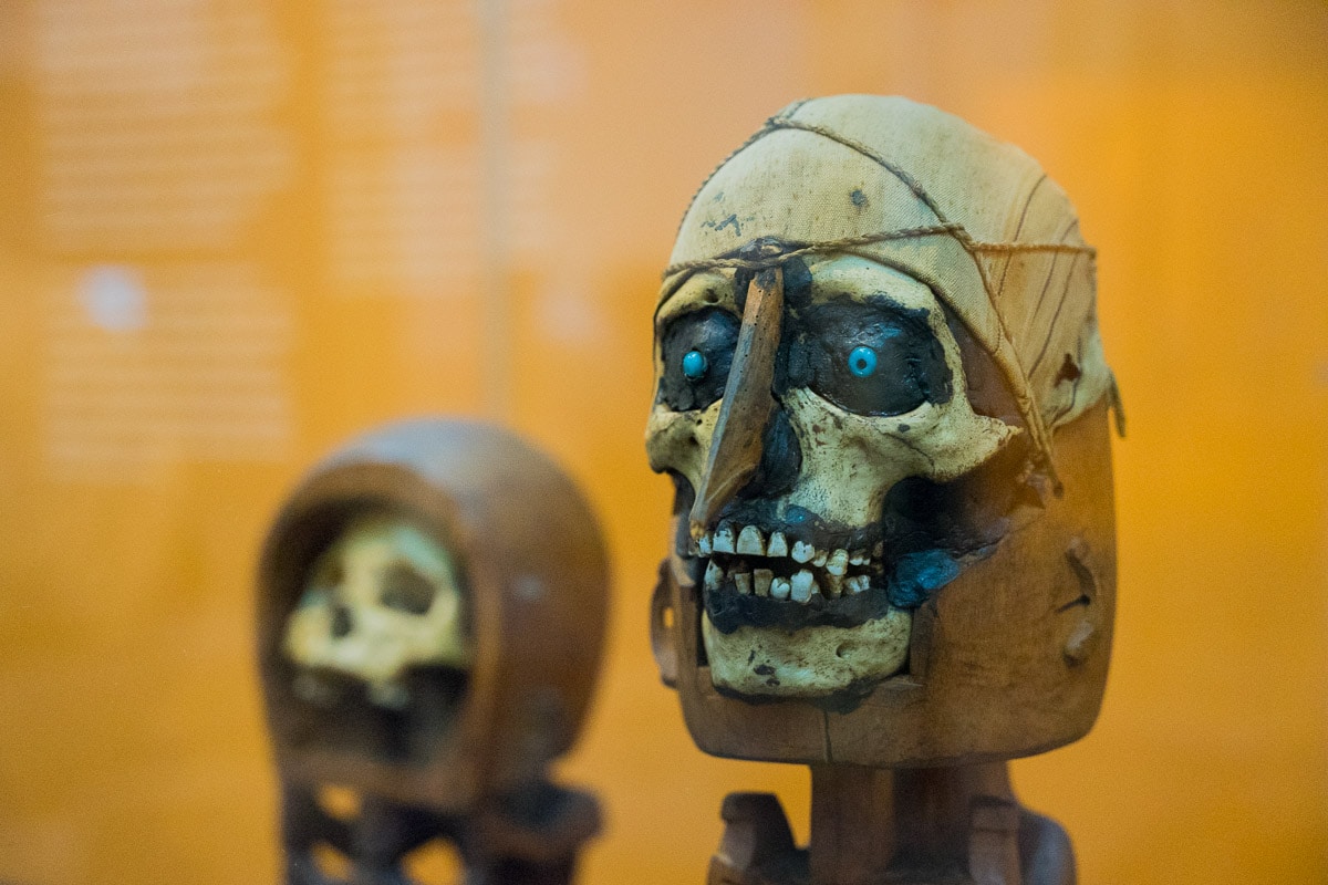 A human skull sitting on top of a wooden statue. There are blue beads in the eye sockets and a wooden nose. This is an artefact from Papua called a korwar and is used to keep the soul of a deceased person