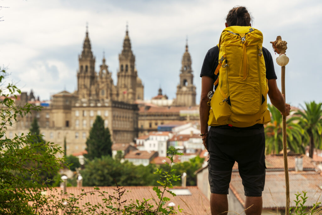 A pilgrim holding a walking stick 
 seen from behind looking at the cathedral of Santiago de Compostela in Spain with a yellow backpack on his back
