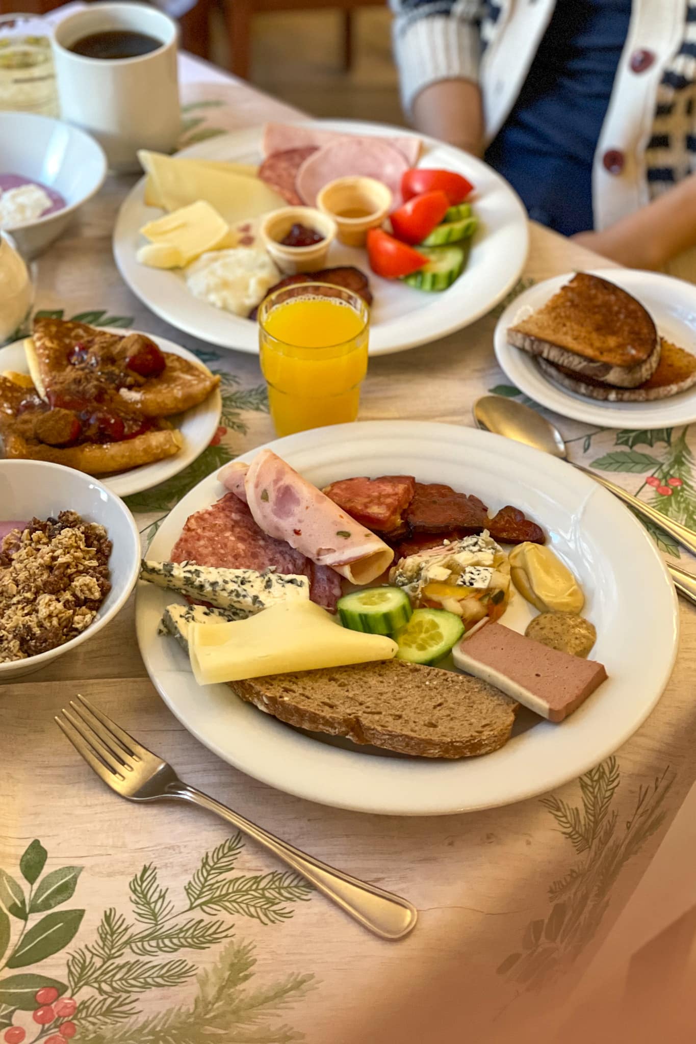 A breakfast plate filled with cold cuts, breads, cheese. There is also orange juice, yoghurt, toast and pancakes with berries