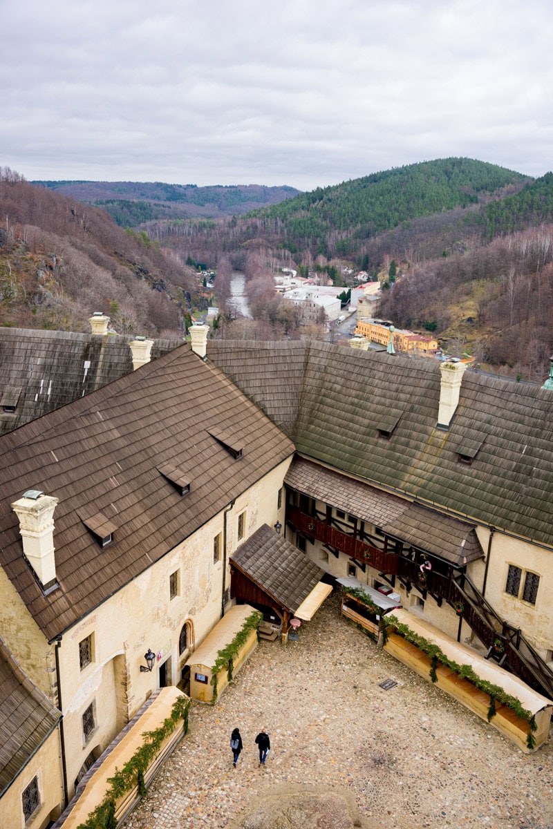 Two visitors walking through the courtyard of the Loket Castle in the Czech Republic, seen from the castle's tower. Beyond the castle buildings, you can see the Ohre River and forested hills