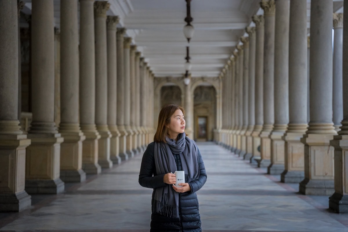 Karlovy Vary in winter: Jin stands between the colomns of the Mill Colonnade holding a traditional spa cup.