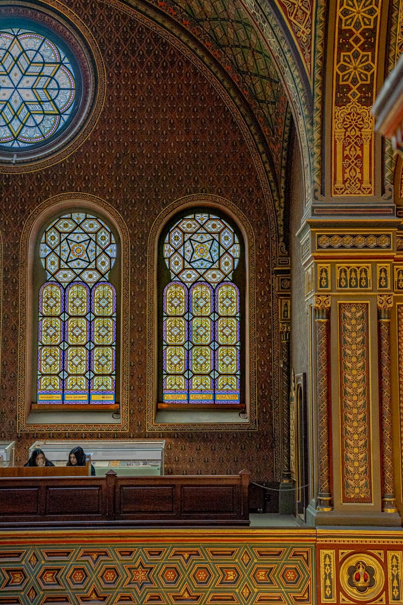 Two women sit on the balcony inside the Spanish Synagogue. They are surrounded by elaborate patterns and colourful stained glass windows