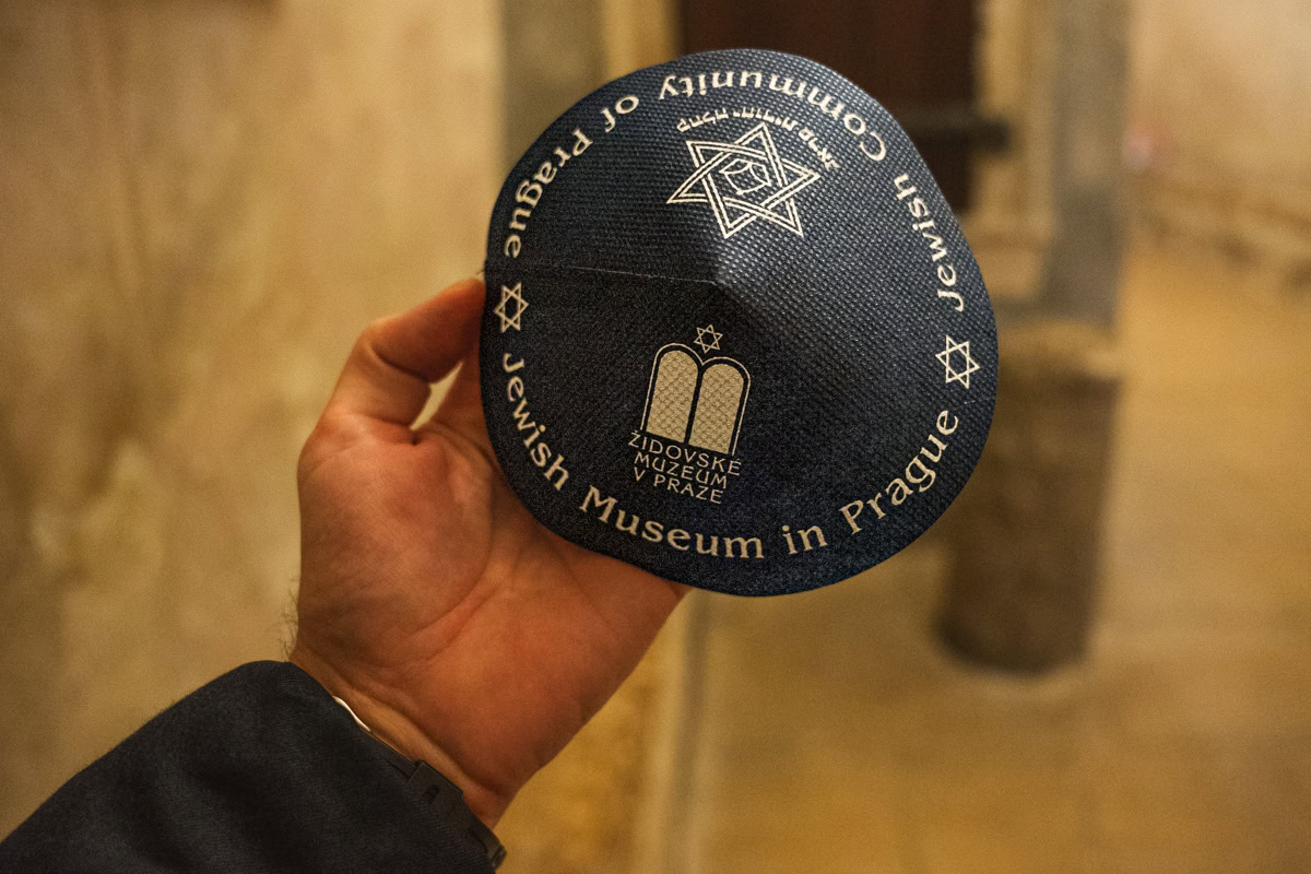 I am holding a kippah (jewish skull cap) in my hand. Men must wear this when entring the synagogues in Prague. It is navy blue with a Star of David on with the phrases Jewish Museum in Prague and Jewish Community of Prague printed on it