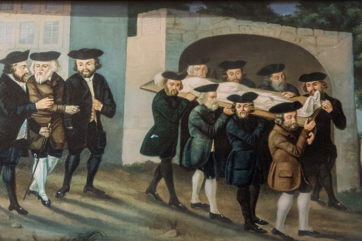 A painting depicting Jewish funeral rites inside the Ceremonial Hall in Prague. Eight bearded men carry a corpse covered in cloth on their shoulders