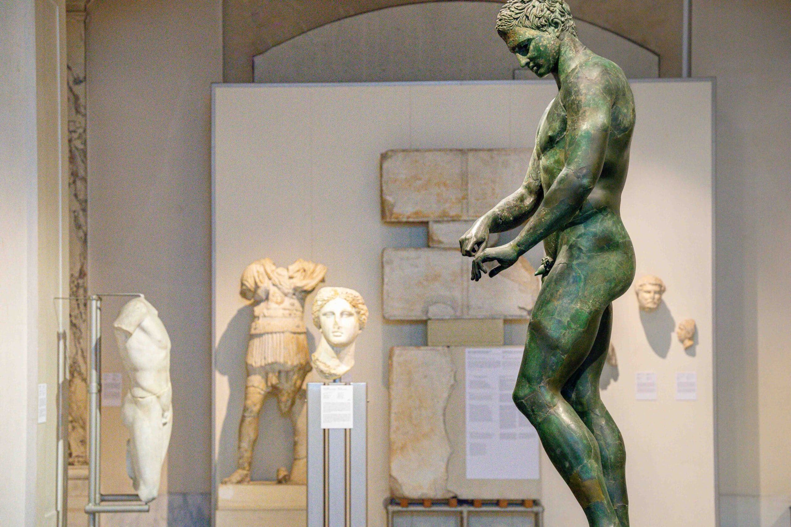 The Bronze Statue of an Athlete, seen in profile with statues in marble behind it. The patina green of the athlete stands in stark contrast to the pale white marble staues behind it