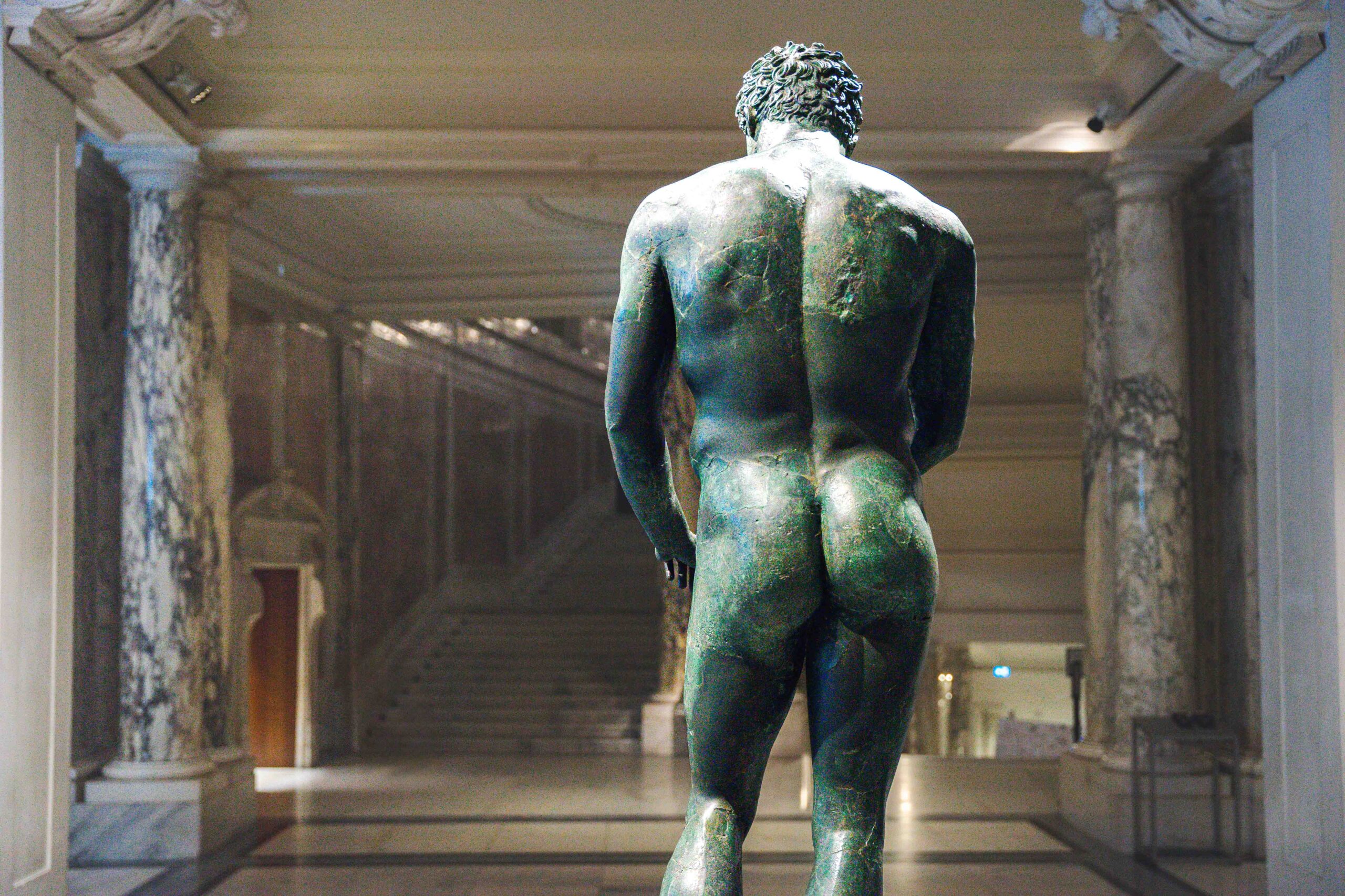 The The bronze Statue of an Athlete in the Ephesus Musem in Vienna, seen from the back. He is naked and this photo shows him from his calves upwards, with his head slightly turned down. In the background you can see marble pillars and stairs of the Neue Burg building, a wing of the Hofburg Palace