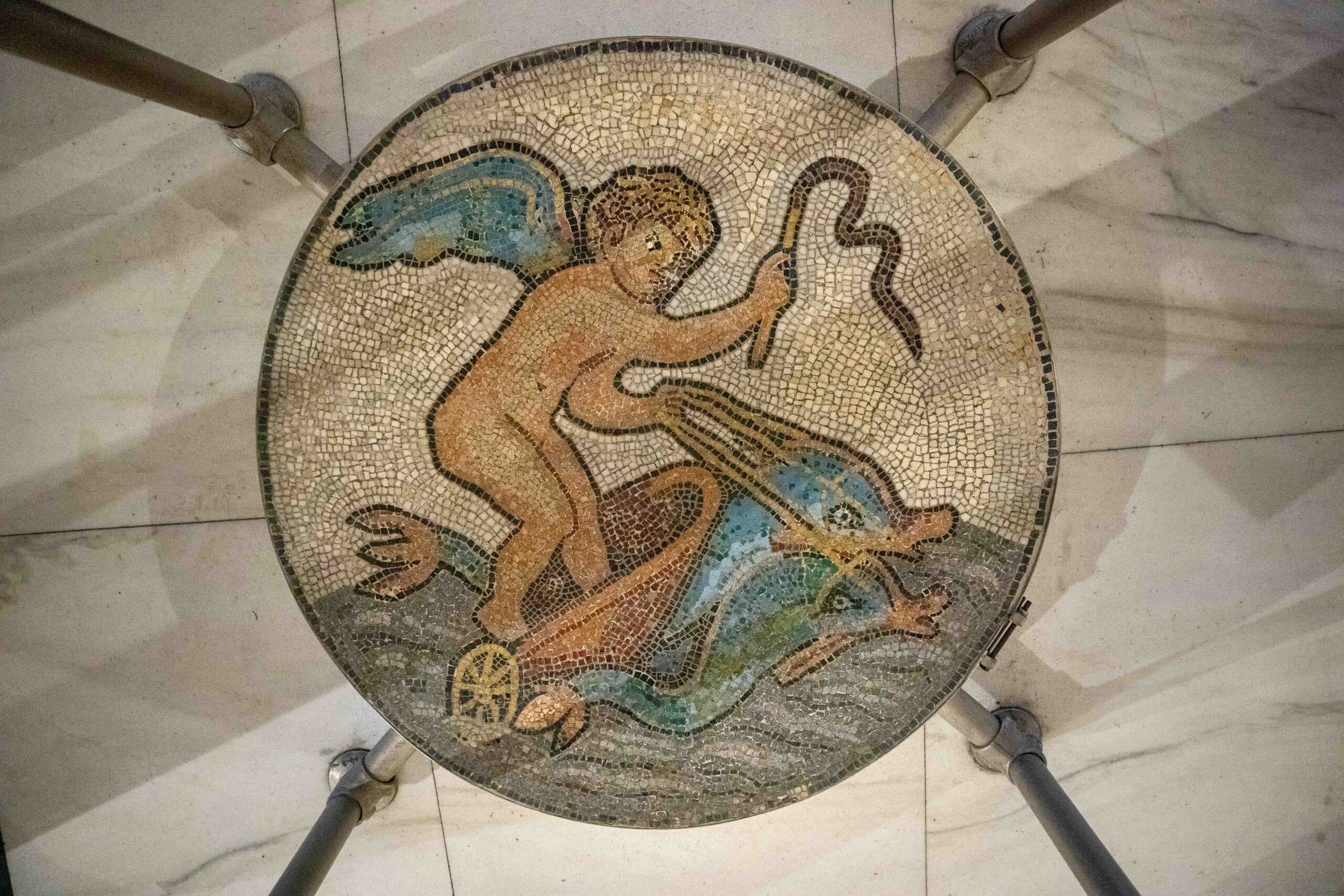 A mosaic in hues of orange, blue, white and gray depicting Cupid riding a chariot drawn by two dolphins
