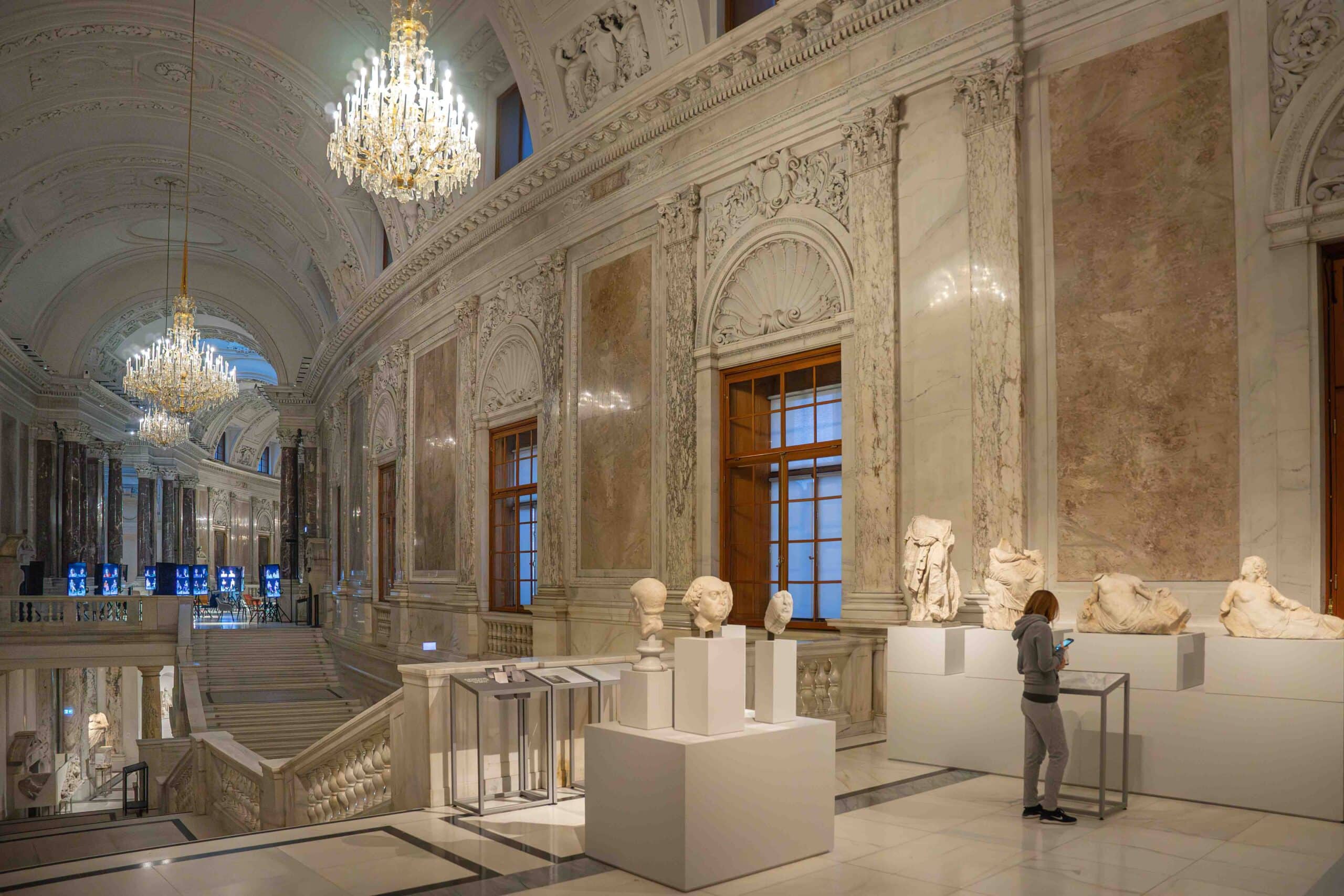 Ephesus Museum Vienna: An interior shot of the Neue Burg, a wing of the Hof Burg Palace. The walls are lavishly decorated with marble and stucco, while chandeliers hang from the barrel shaped ceiling. In the foreground, you can see my wife reading information between displays of Roman statues and busts in marble