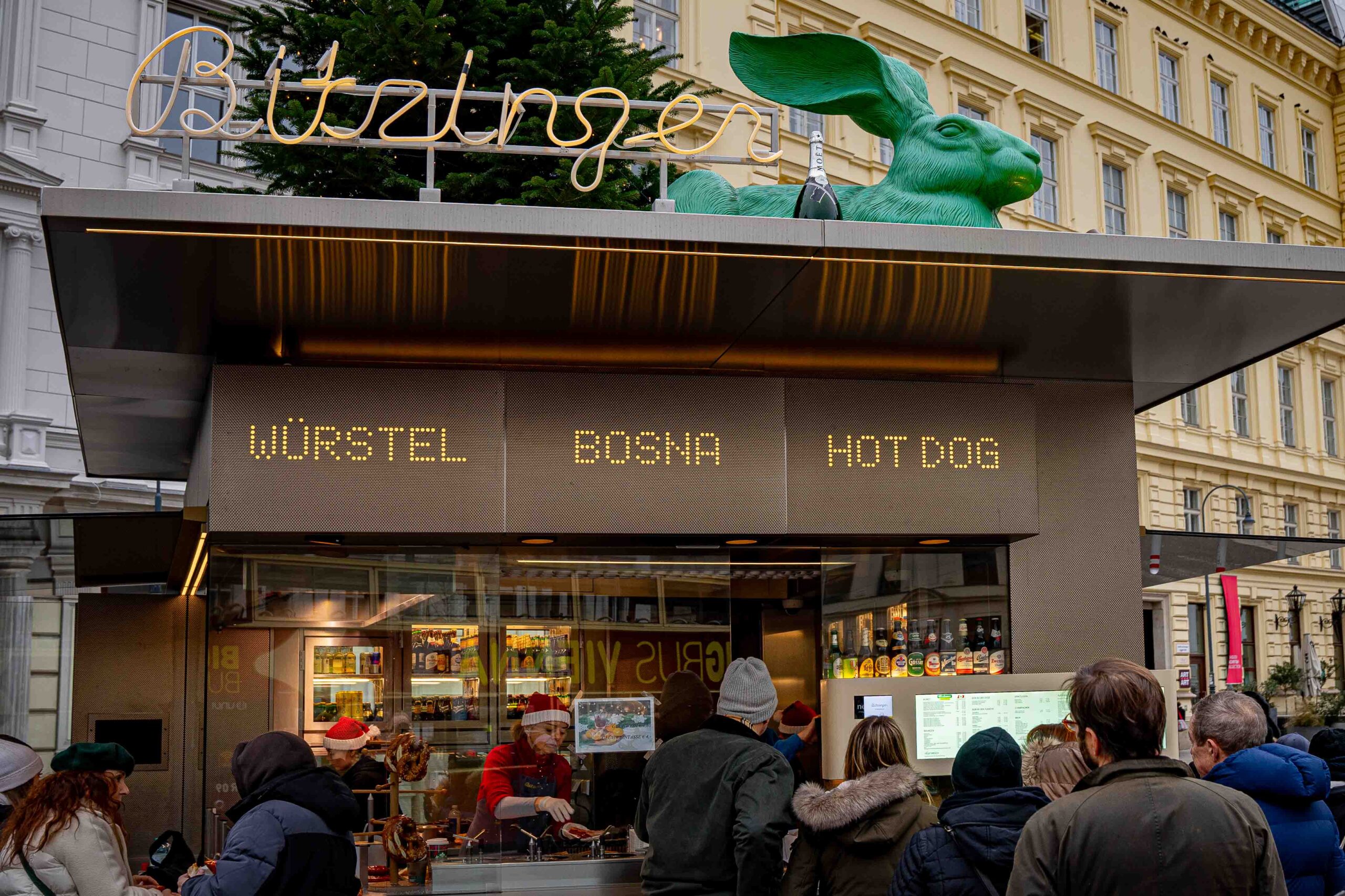 Bitzinger Wurstel stand in Viennna. You can see the words wurstel, bosna and hot dog displayed on the stand. On the roof the iconic green rabbit sits behind a bottle of Moet champagne