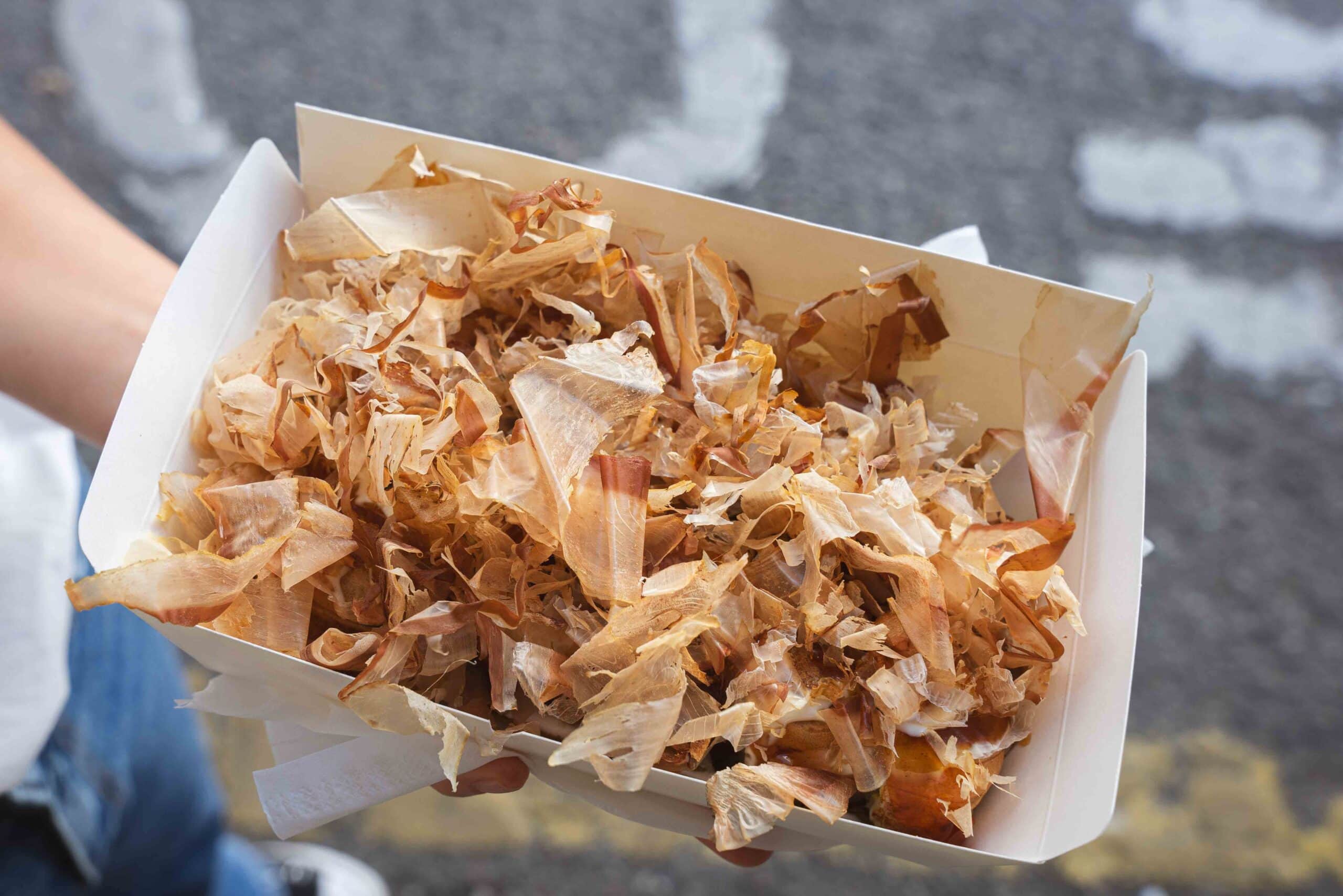  Jin is holding a paper box with a baffle from the Netflix food stand in Dongdaemun.It's entire covered by moving bonito fish  flakes 