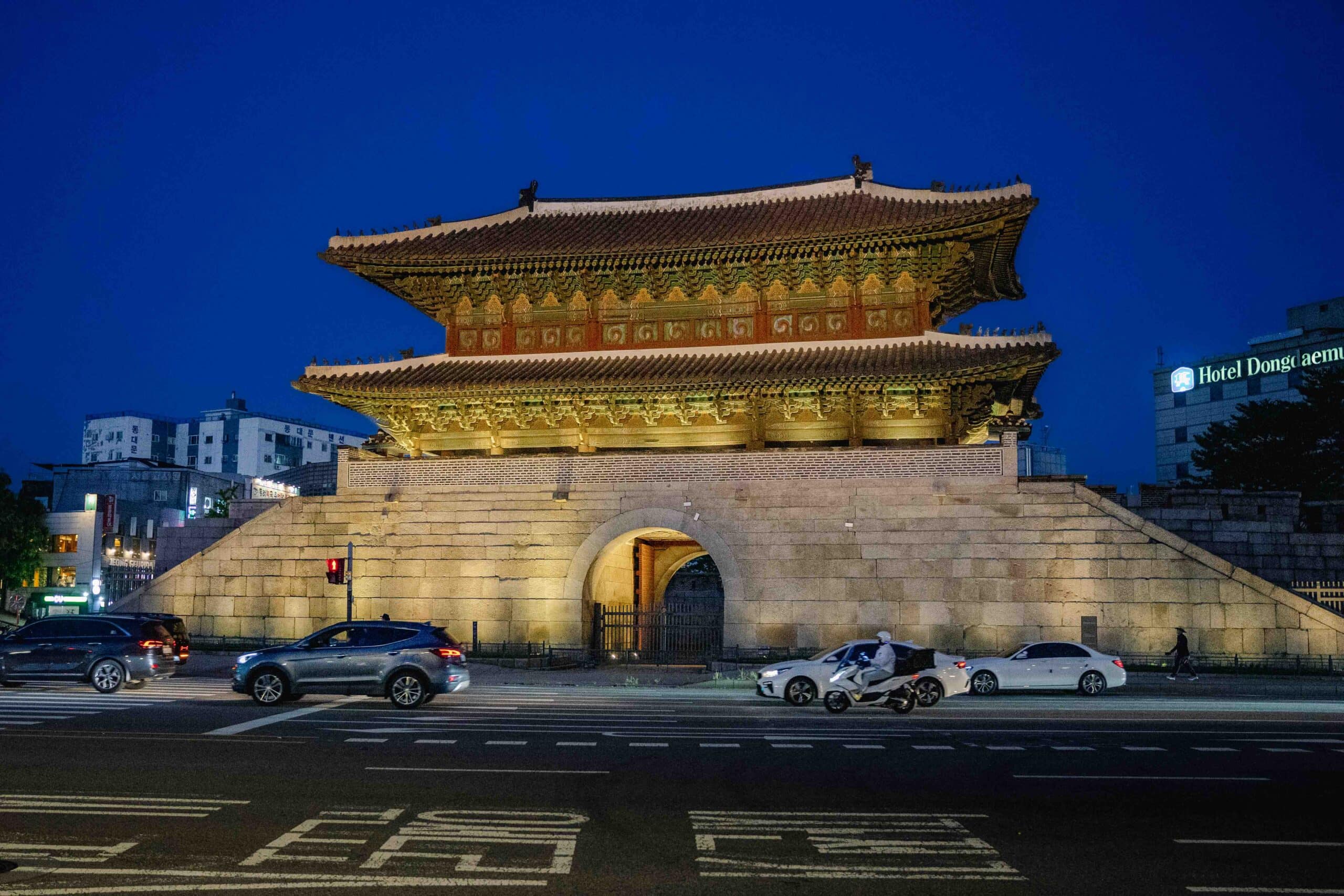 Heunginjimun (Dongdaemun Gate) lit up at night with traffic driving by. Behind the gate you can see the Hotel Dongdaemun