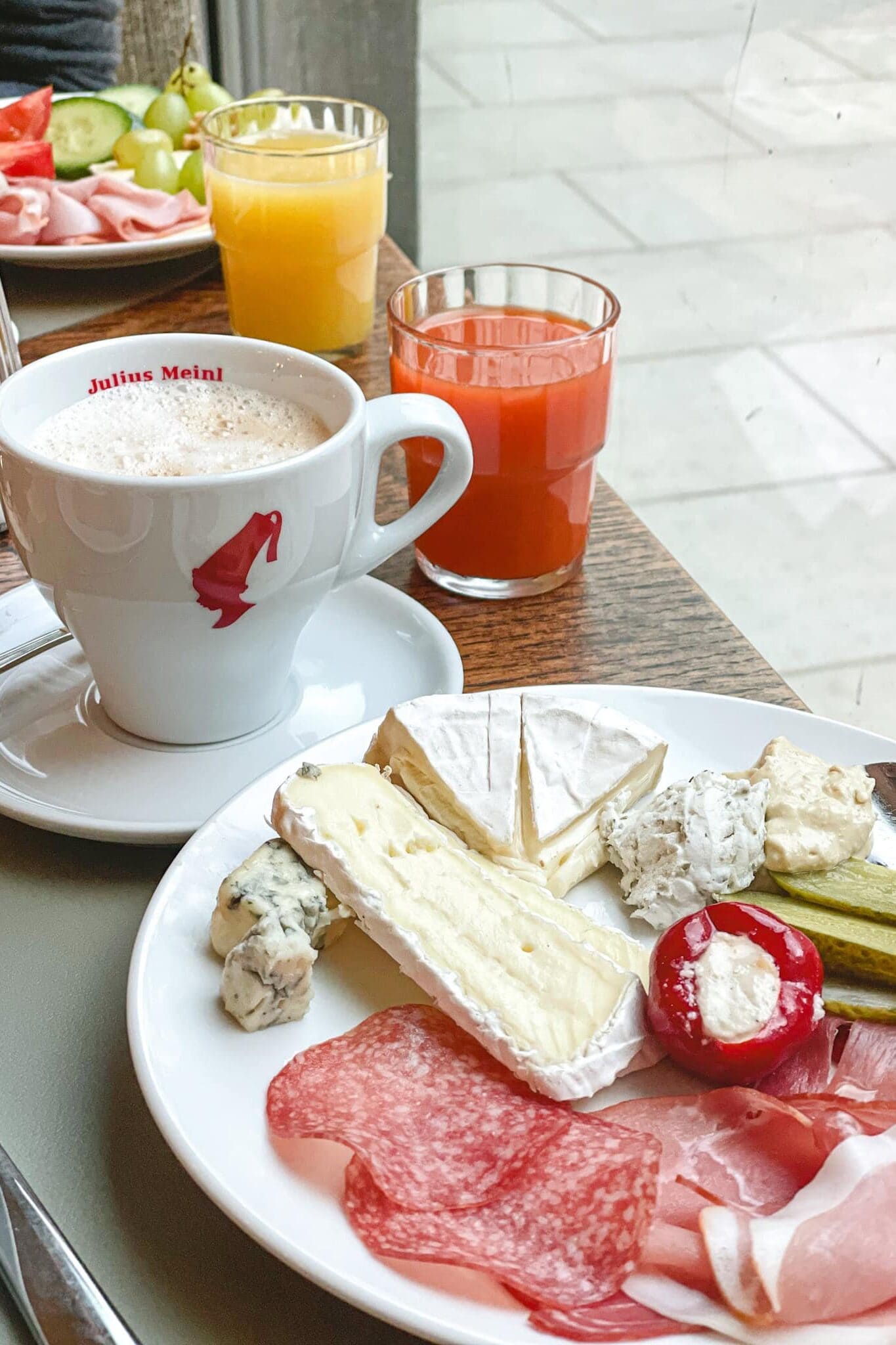 My breakfast plate at Weekend Hotel in Vienna. The plate is full of various cheeses, salami, ham and gherkins. There is also Julius Meinl coffee and two glasses of juice on the table
