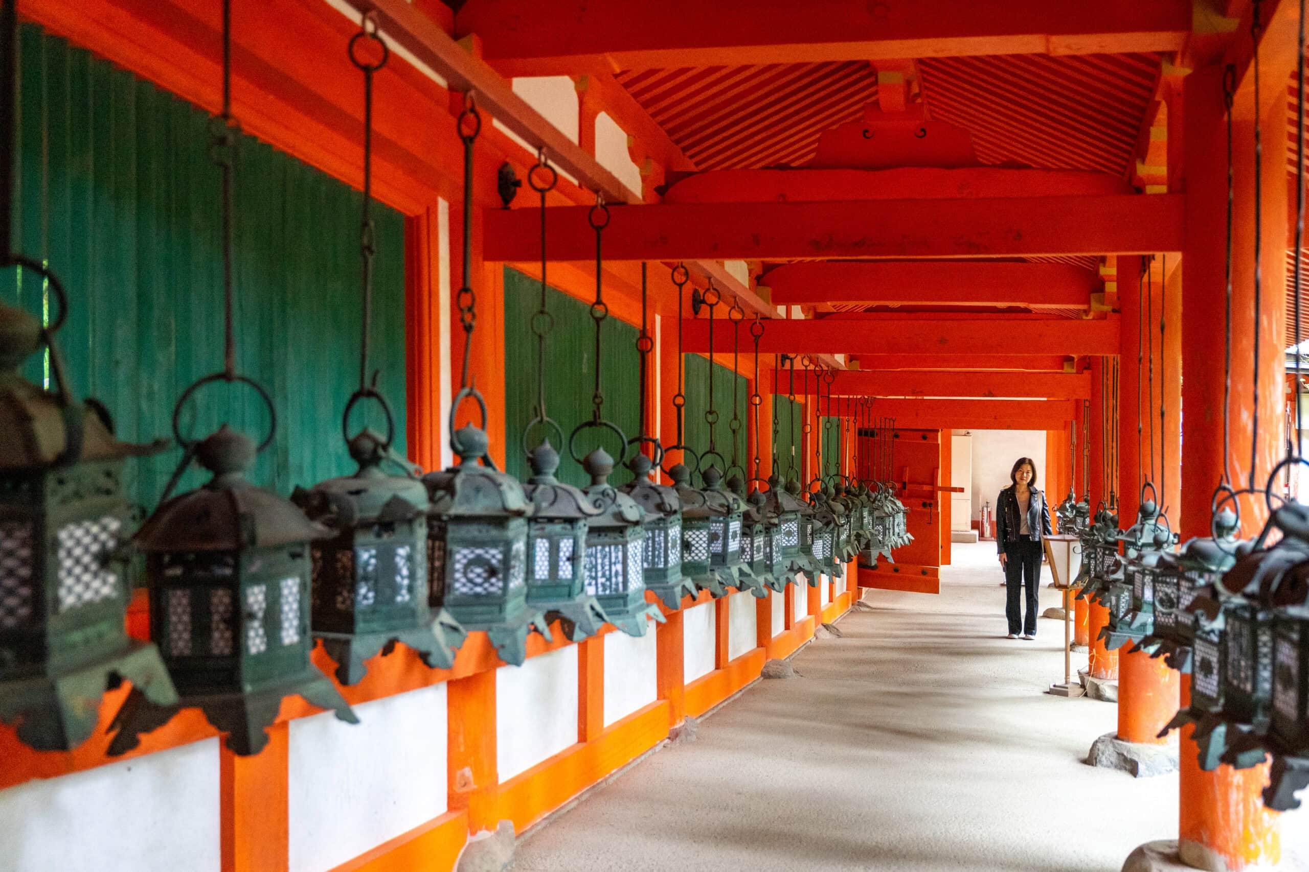 Jin stand at the end of a red and green corridor lined with bronze lanterns at the Kasuga Taisha Shrine in Nara