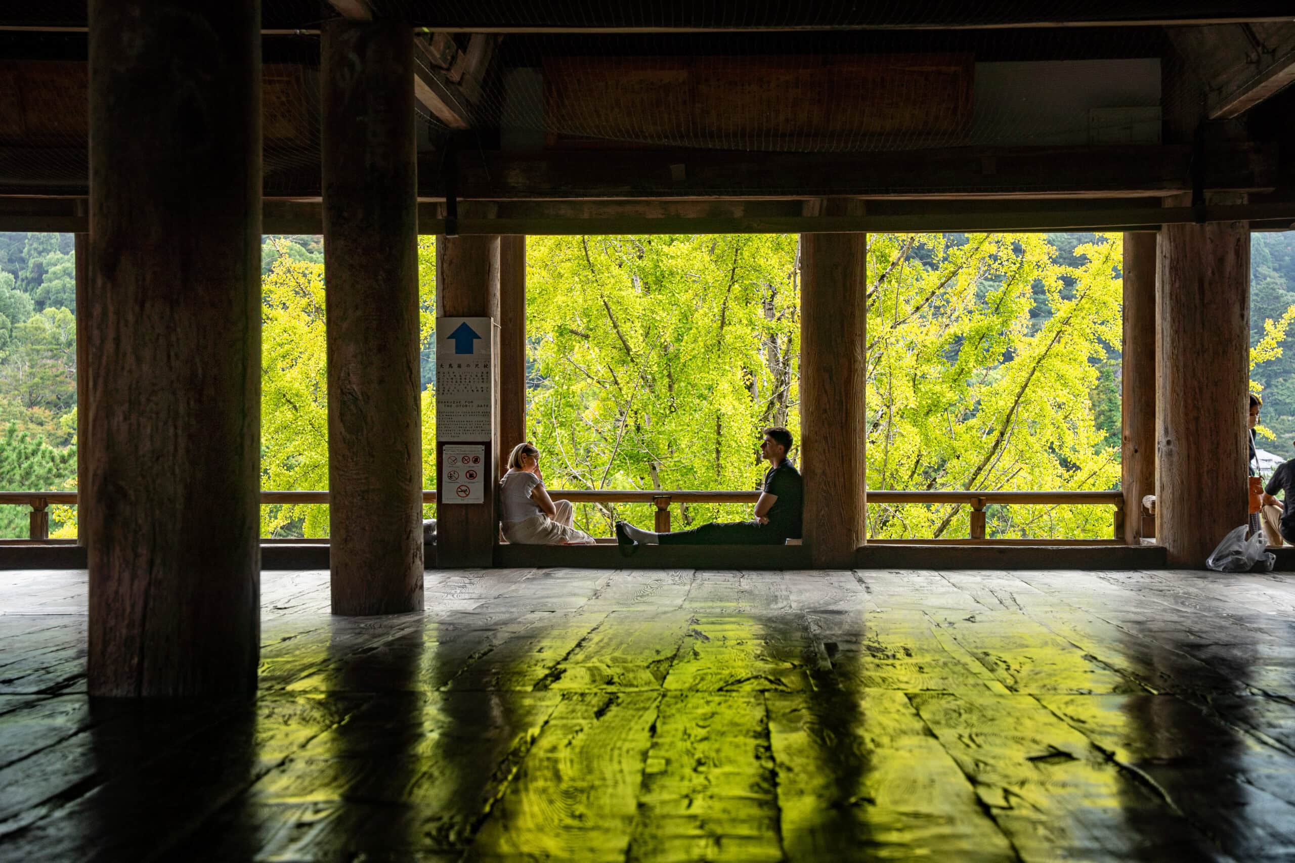 A wester couple sits on the wooden floor with yellow-green leaves behind them inside the Hokoku Shrine on Miyajima island