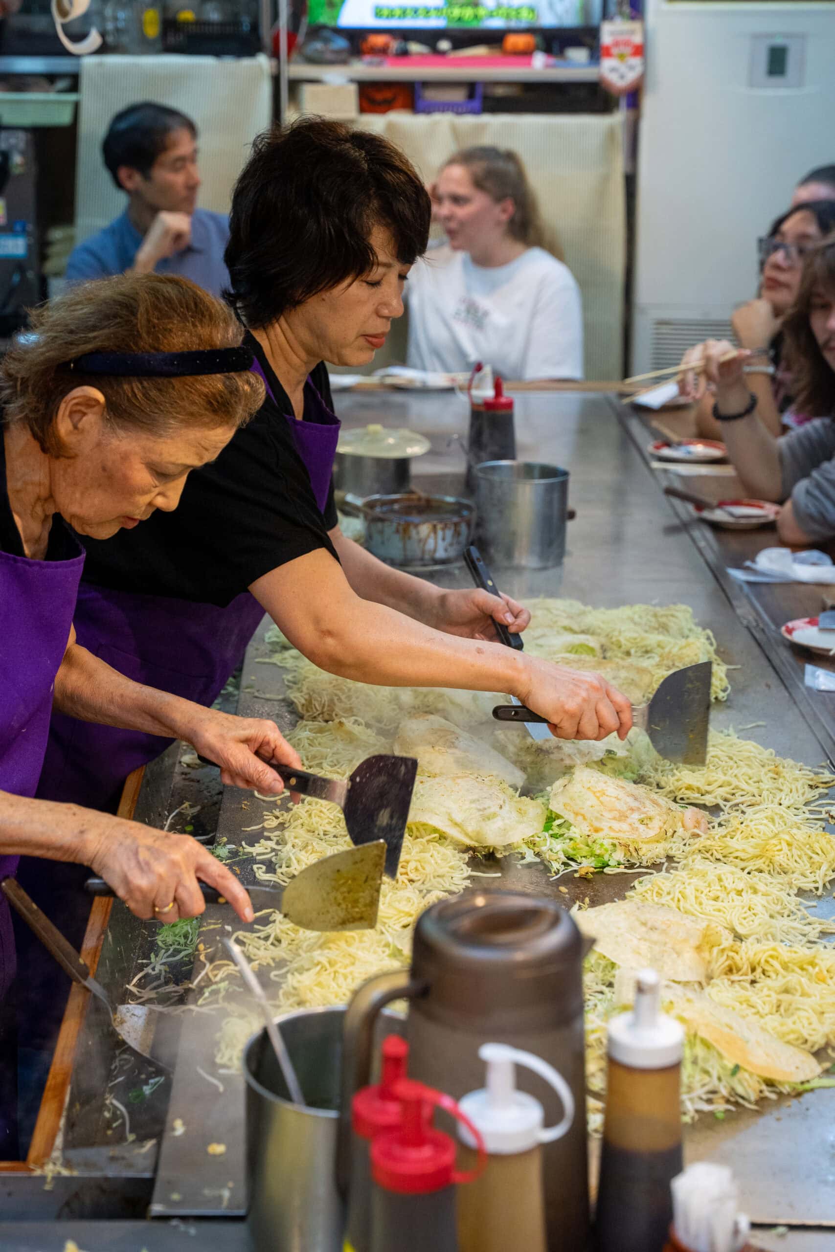 Two Japanese women prepare okonomiyaki at Okonomimuran Hiroshima while western tourist look and and wait for their food