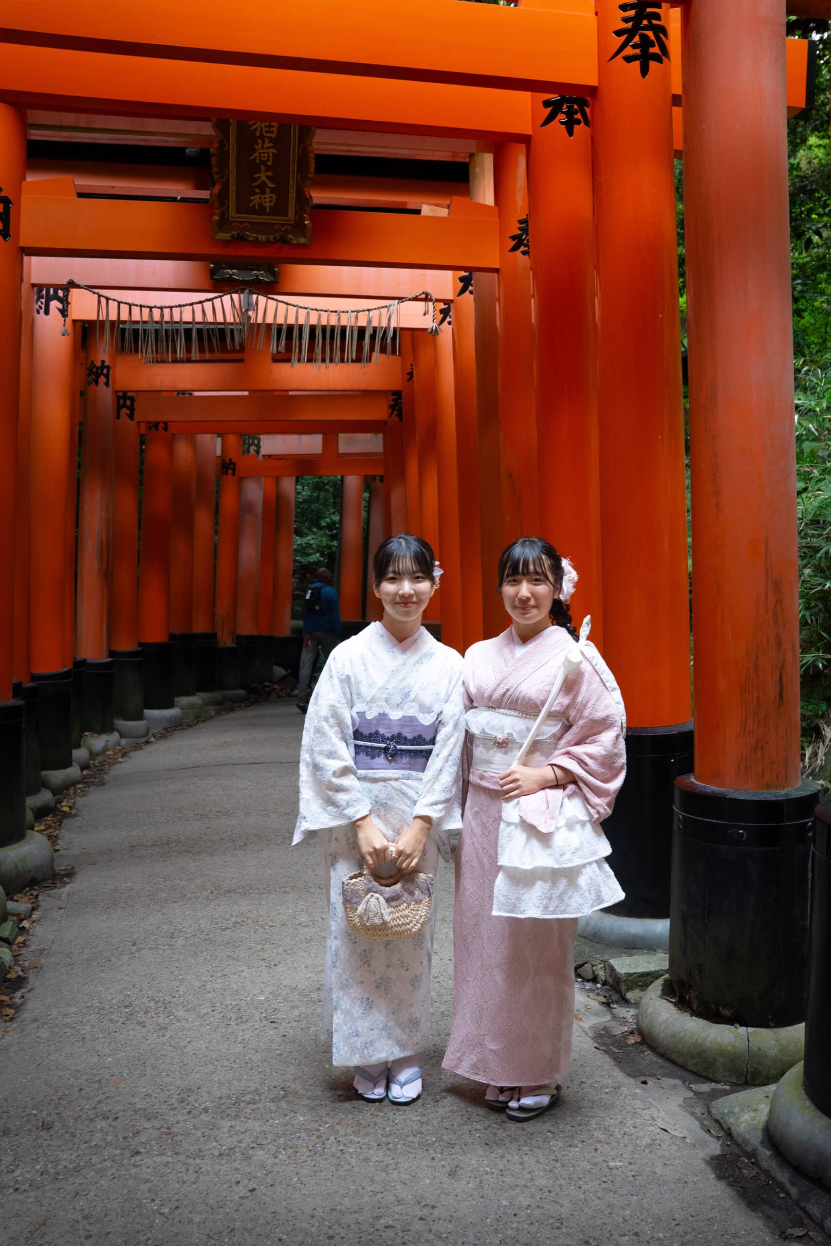 two asian girls wearing traditional Japanese dress stand under the ornage torii gates of Fushimi Inari near Kyoto, Japan