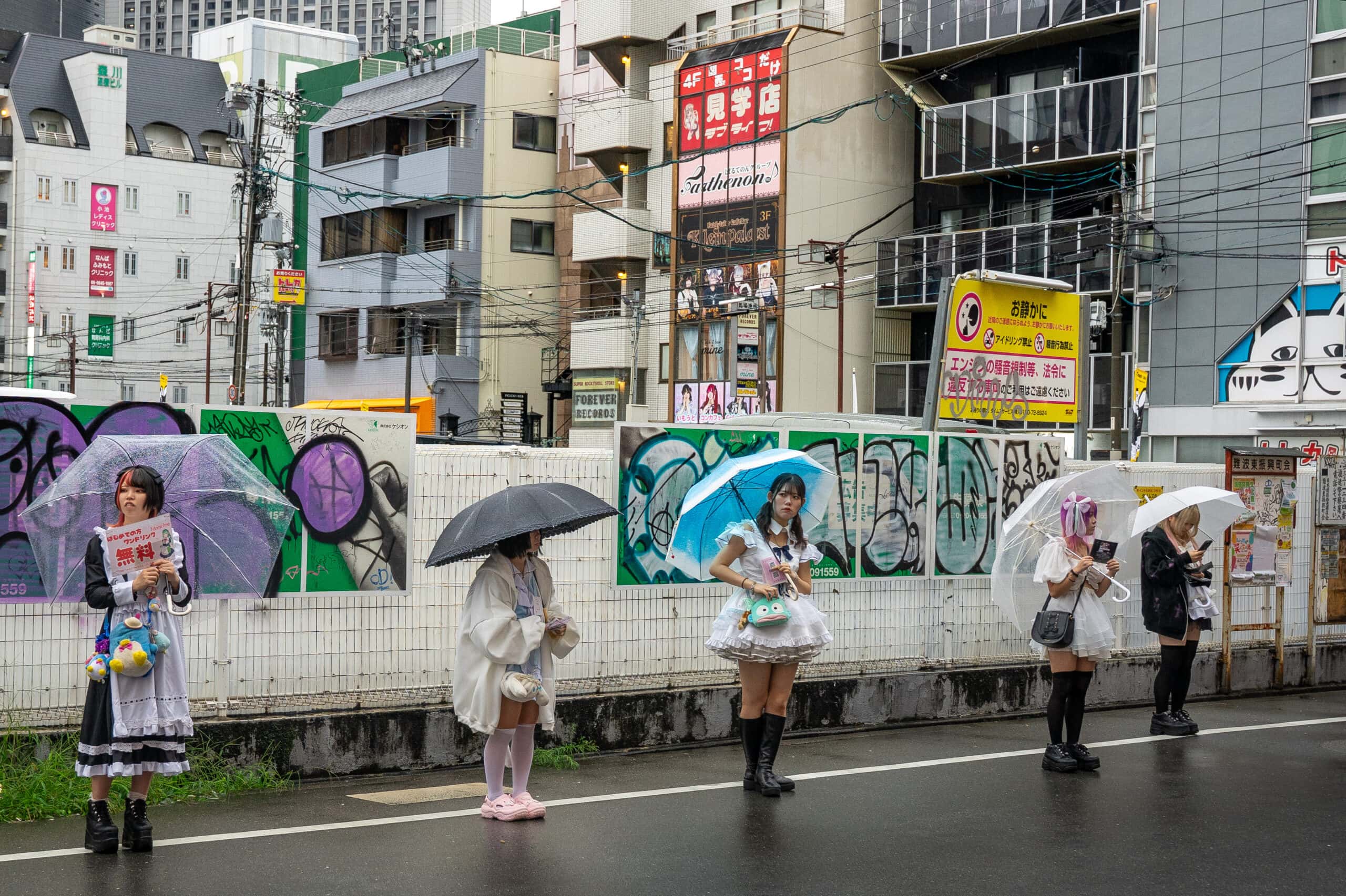 Maids wearing frilly costumes stand under umbrellas in the rain, seen on the street in Osaka. They are waitresses in maid cafes