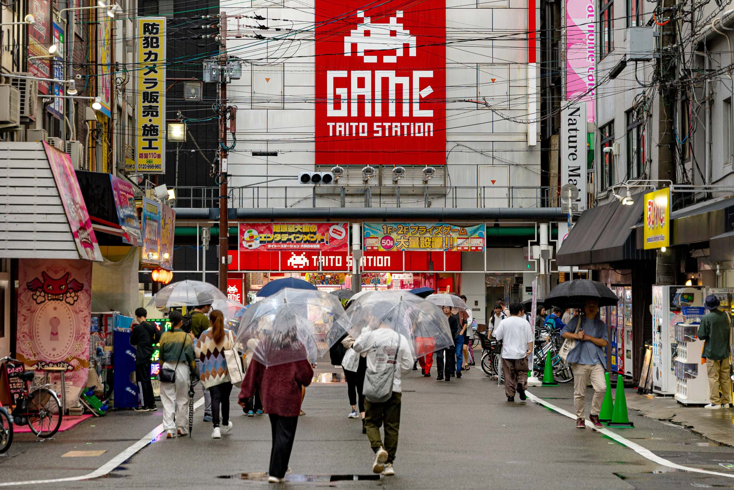 People walk under umbrellas towards a building with a red sign saying GAME TAITO STATION in Osaka