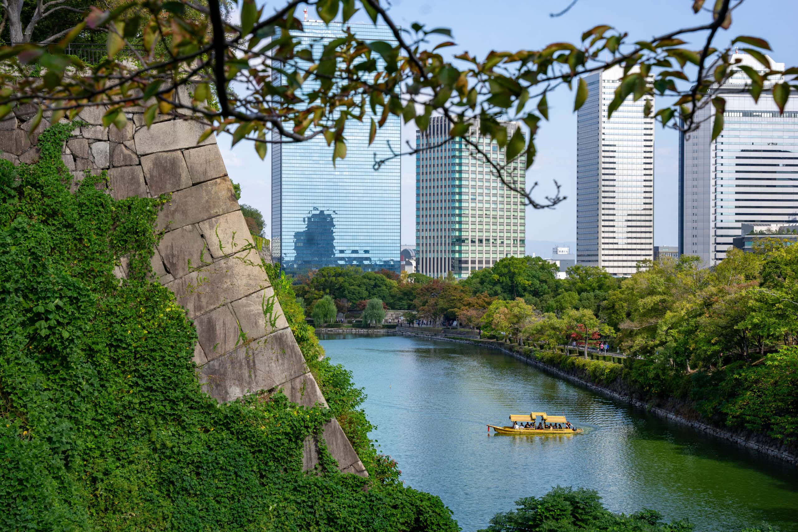 2 days in Osaka itinerary: a yellow boat takes visitors on a cruise along the moat of Osaka Castle. You can see modern skyscrapers in the back ground with a section of the castle's stone wall in the foreground