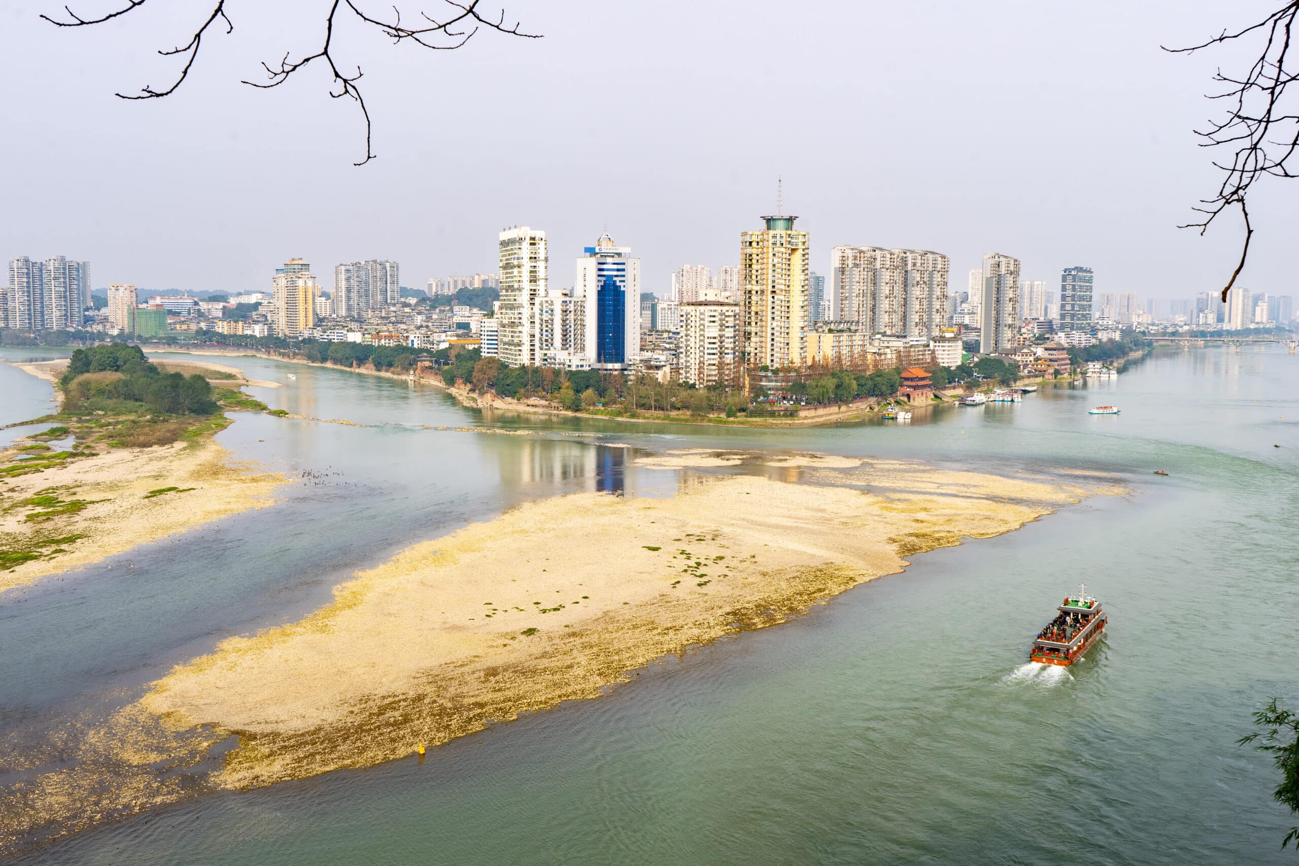 A scene of a tier3 city in China. There is a river with a boat and tall buildings on the river's edge