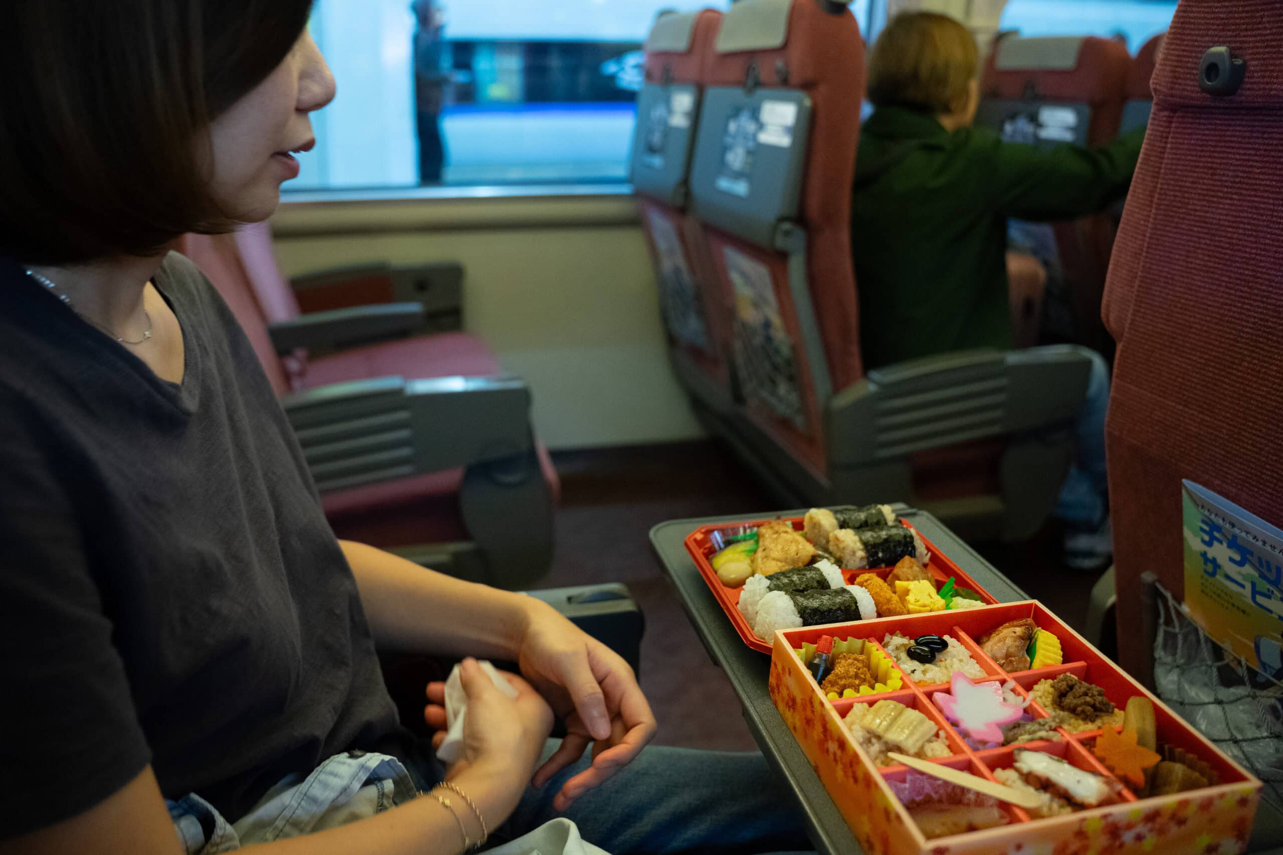 Jin sitting in a seat on a train from Hiroshima to Kinosaki Onsen with two bento boxes