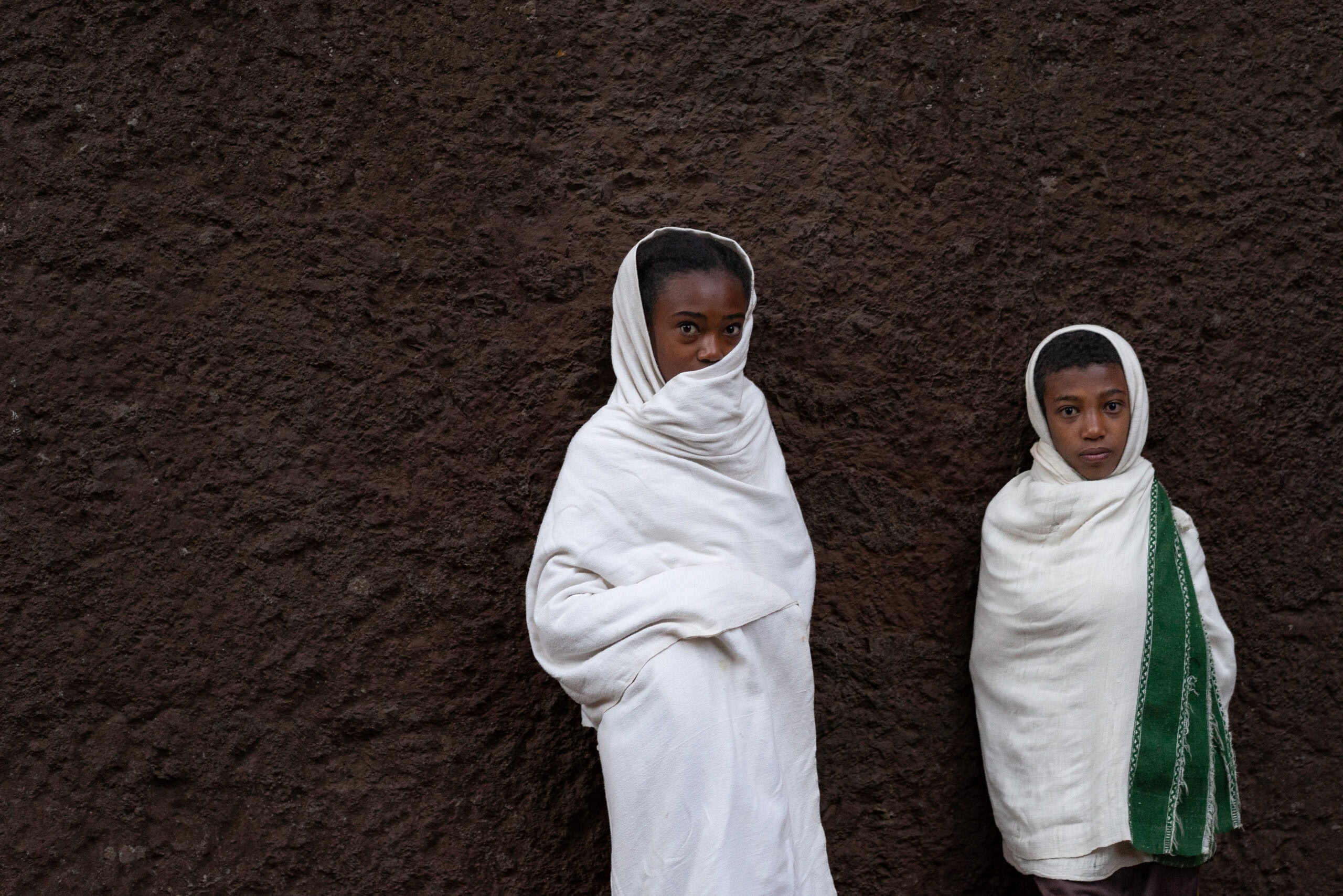 two children wrapped in white shawls stand agains the wall of a rock hewn church in Lalibela, Ethiopia