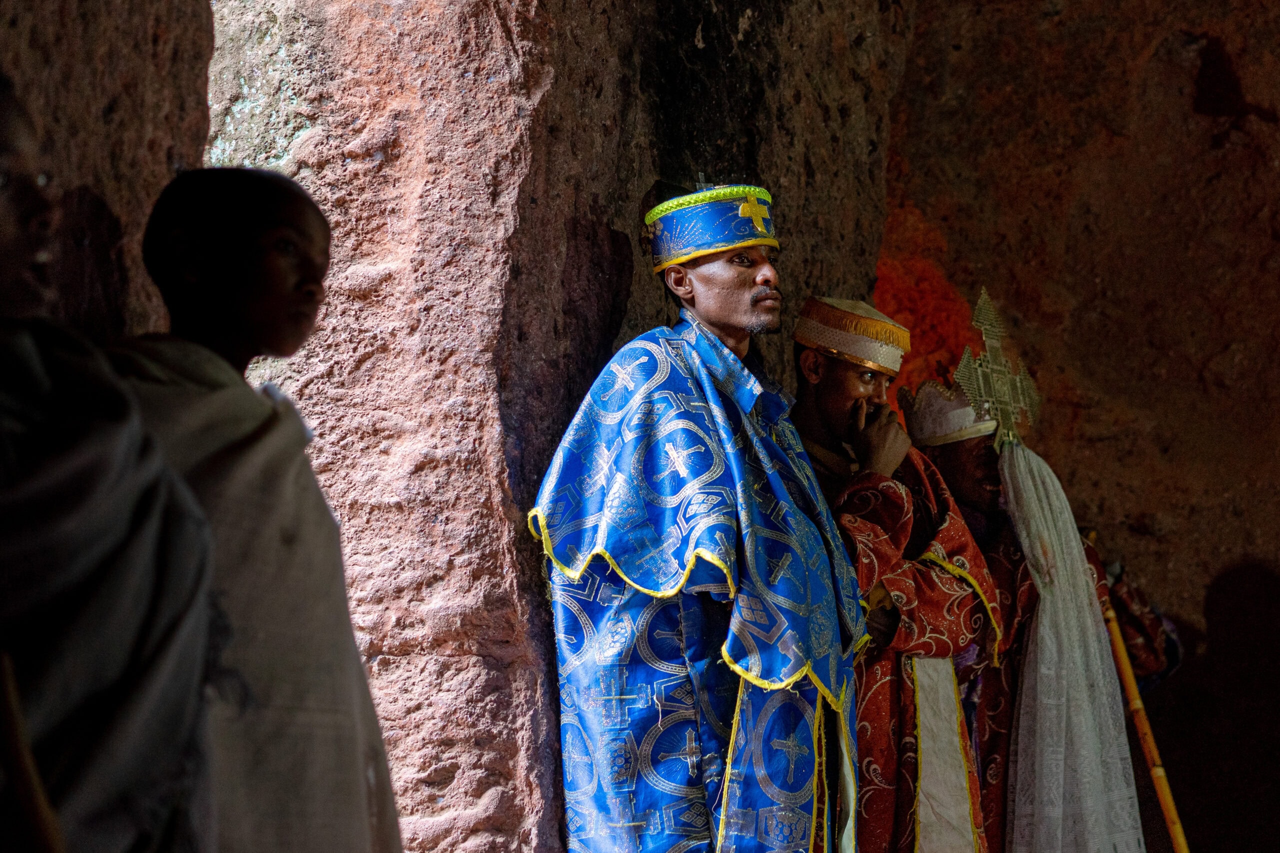 Three church deacons dressed in blue and crimson stand against a church wall as they listen to Sunday mass inside Biet Giyorgis, the church of Saint George, Lalibela