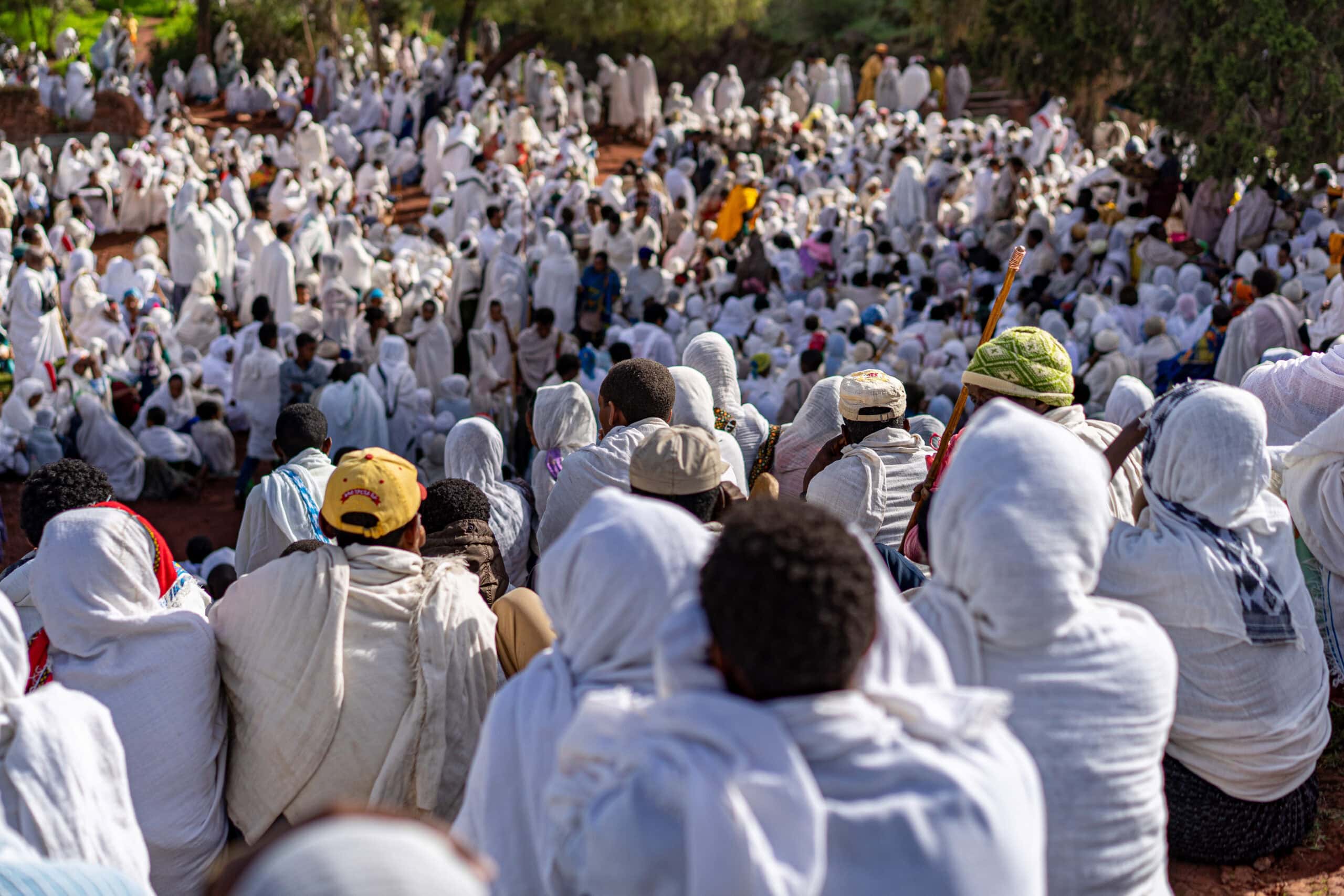A large group of worshippers dressed in white shawls sit outside. There are hundreds of them