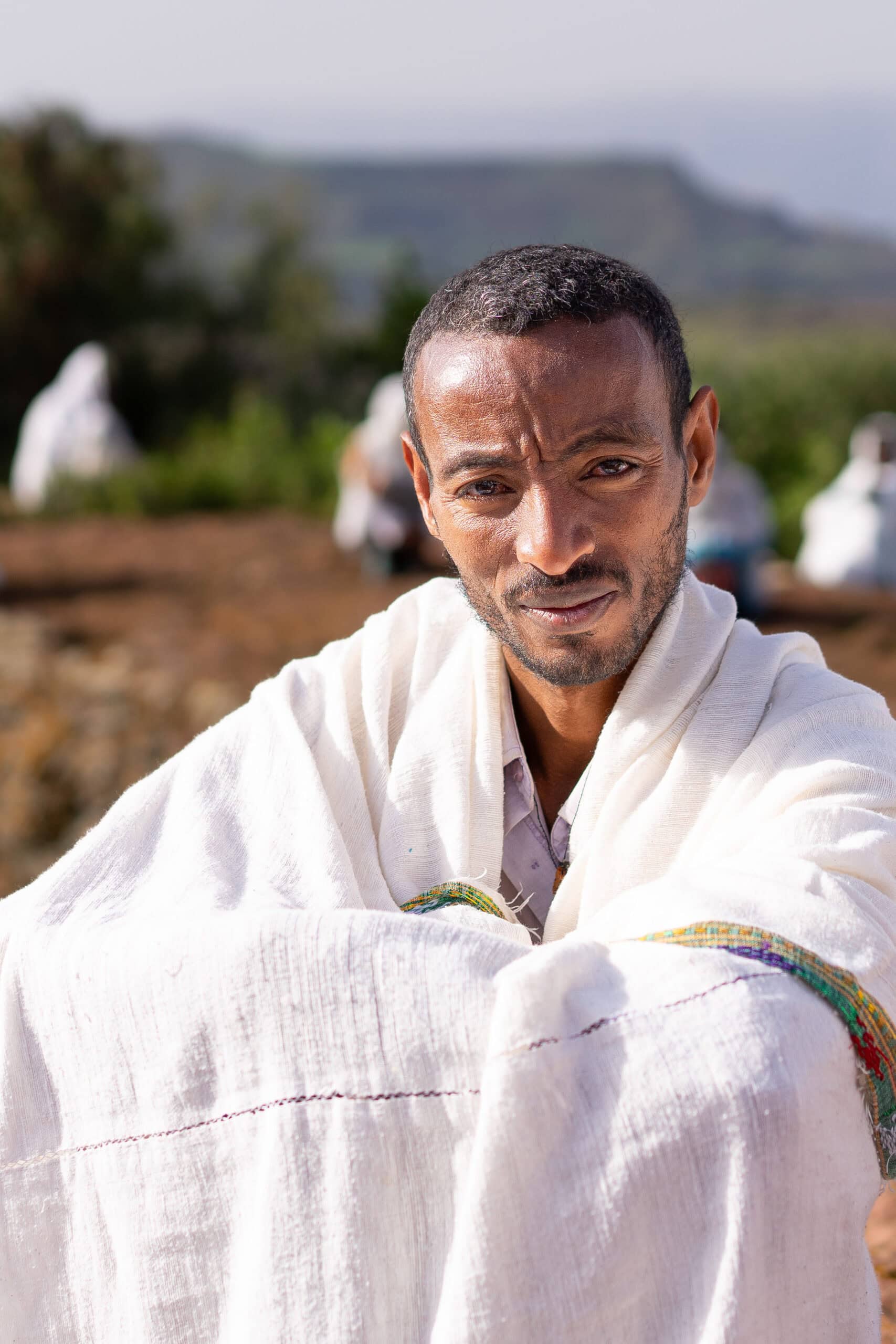 A man with a deep expression, dressed in a white shawl sits outside in the sun on a Sunday morning in Lalibela, Ethiopia