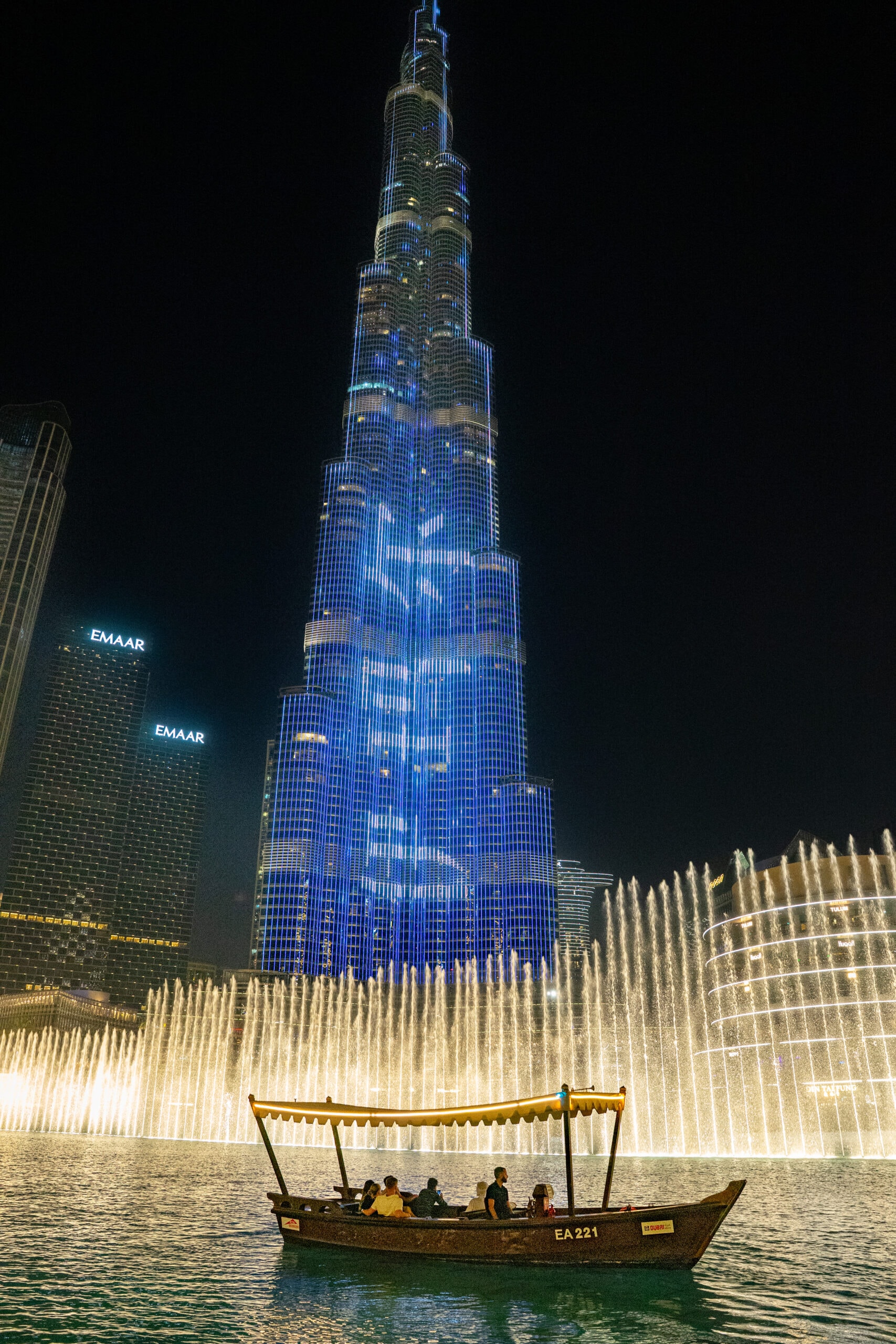 An abra floats in front of the Dubai fountain at night with the Burj Khalifa towering behind